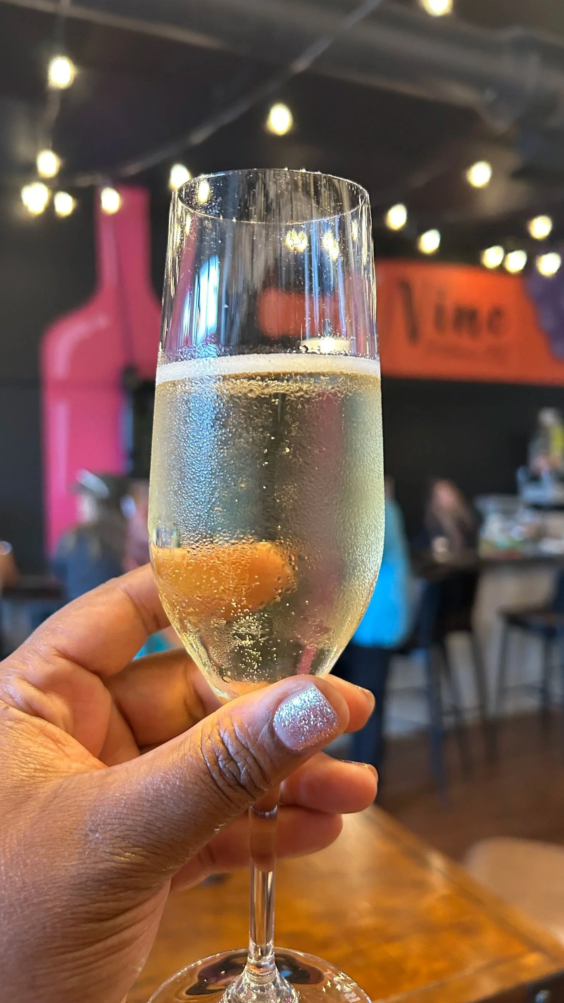 A hand holding a champagne flute filled with sparkling wine in a dimly lit Vino wine bar with people in the background and string lights hanging from the ceiling.