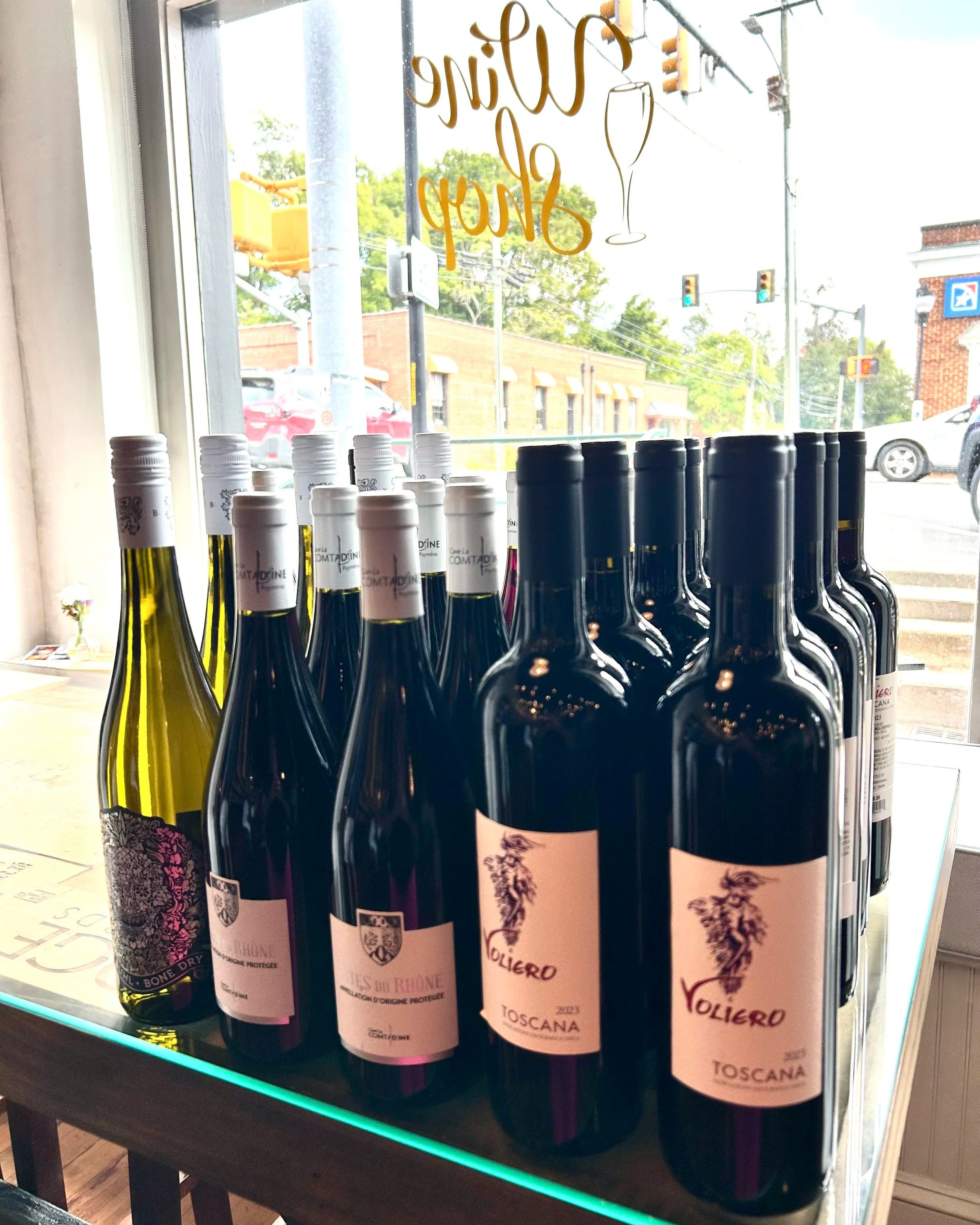 Display of multiple bottles of wine arranged on a table inside a wine shop, with a large window showing a street scene outside.