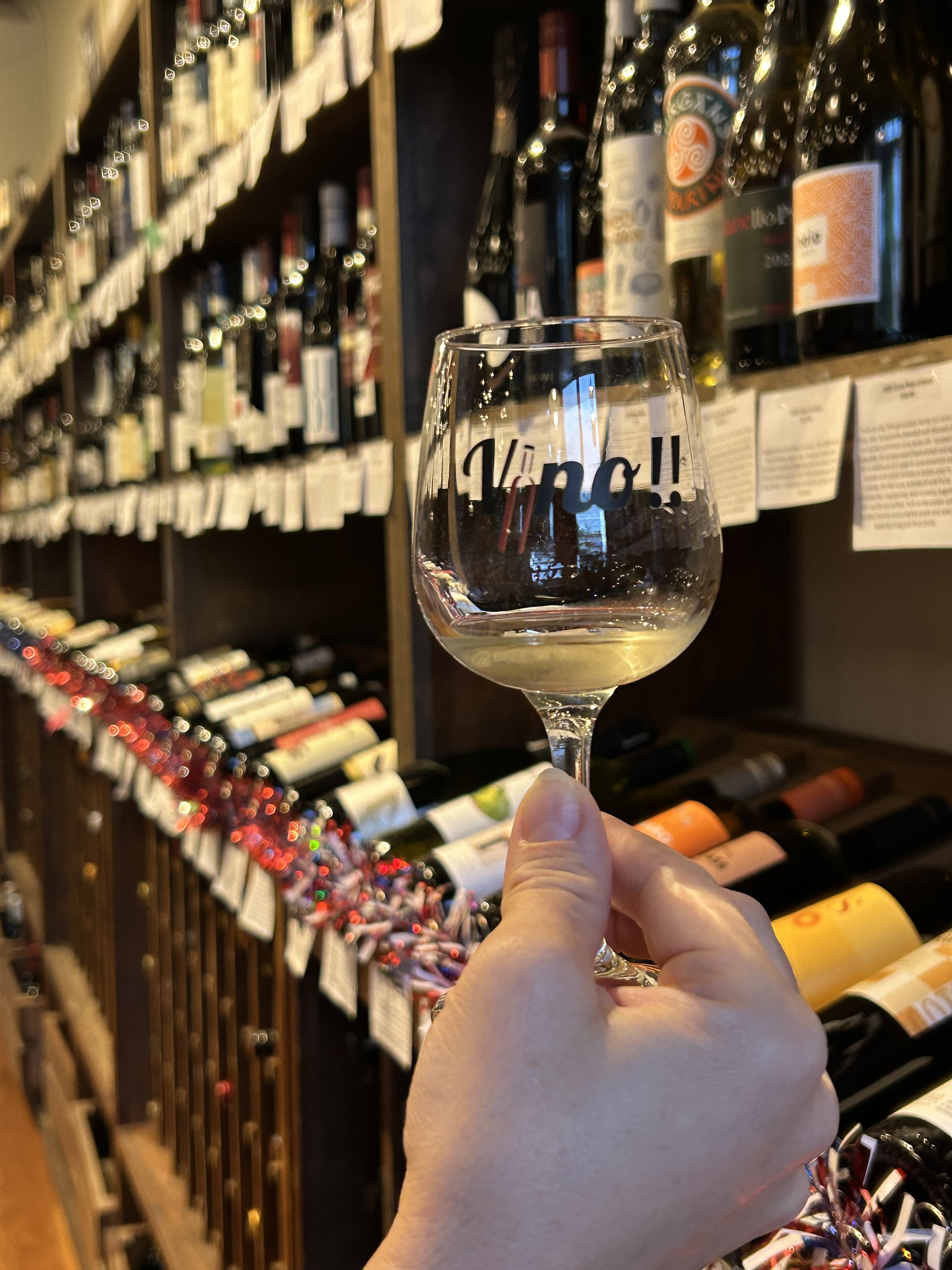 A hand holding a wine glass with the word 'vino!!' on it, in front of a wine shelf filled with various bottles.