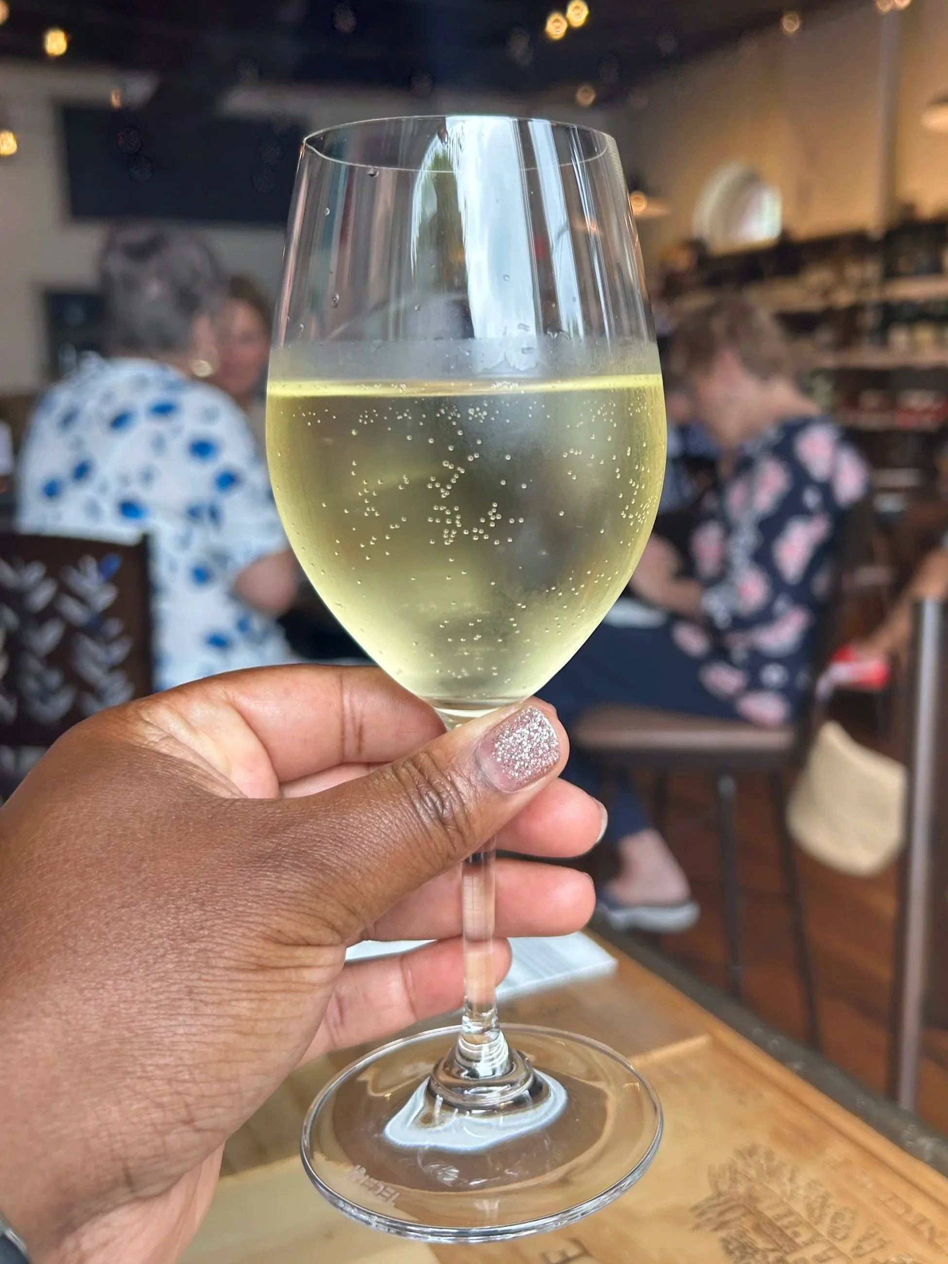 Hand holding a glass of white wine in a restaurant with people sitting and talking in the background.