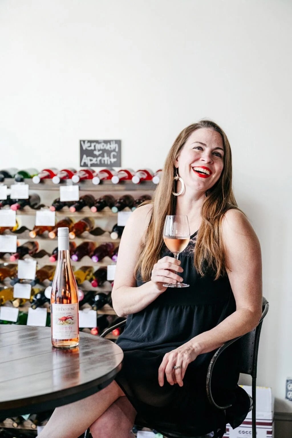 A woman with long brown hair, wearing a black dress and large earrings, sitting at a round table while holding a glass of rosé wine, smiling, with a wine rack in the background labeled 'Vermouth Aperitif'.