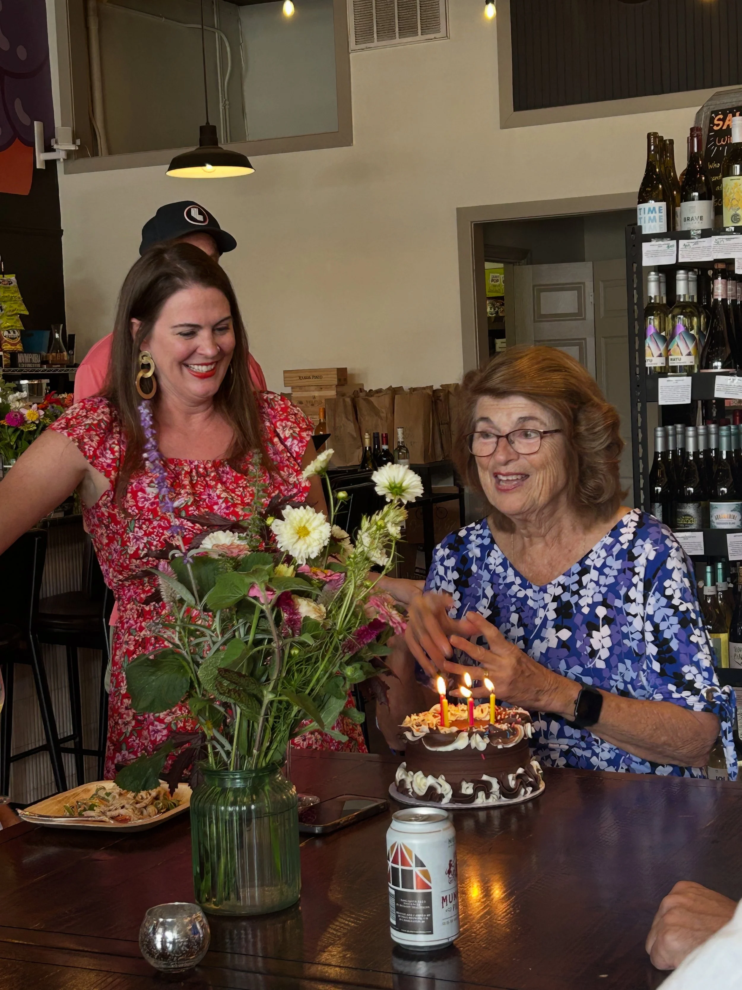 Woman with glasses and short hair celebrating birthday with a cake with lit candles, flowers in a vase, and a can on the table, while other women smile and watch.