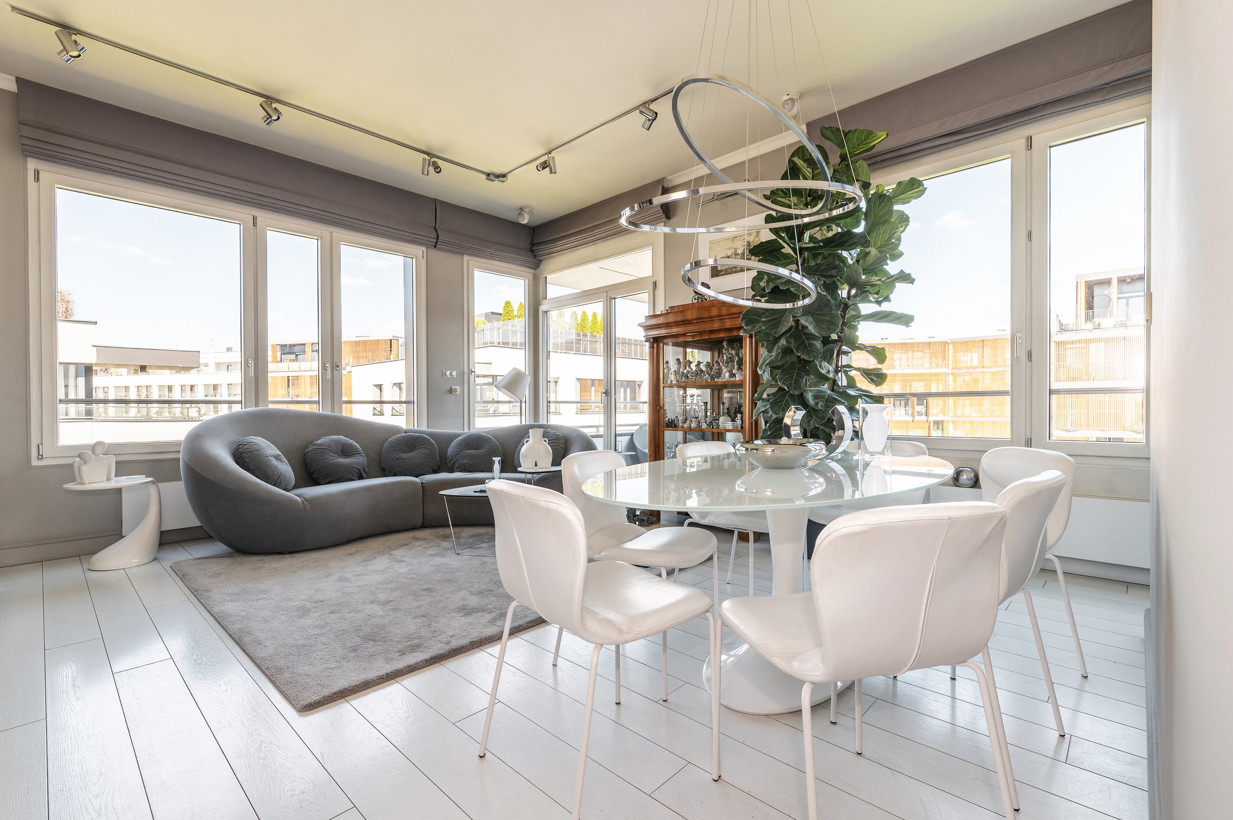 Bright living room with large windows, white dining table with six chairs, a curved gray sofa with pillows, a wooden cabinet with decorative items, a large plant, and modern lighting fixtures.