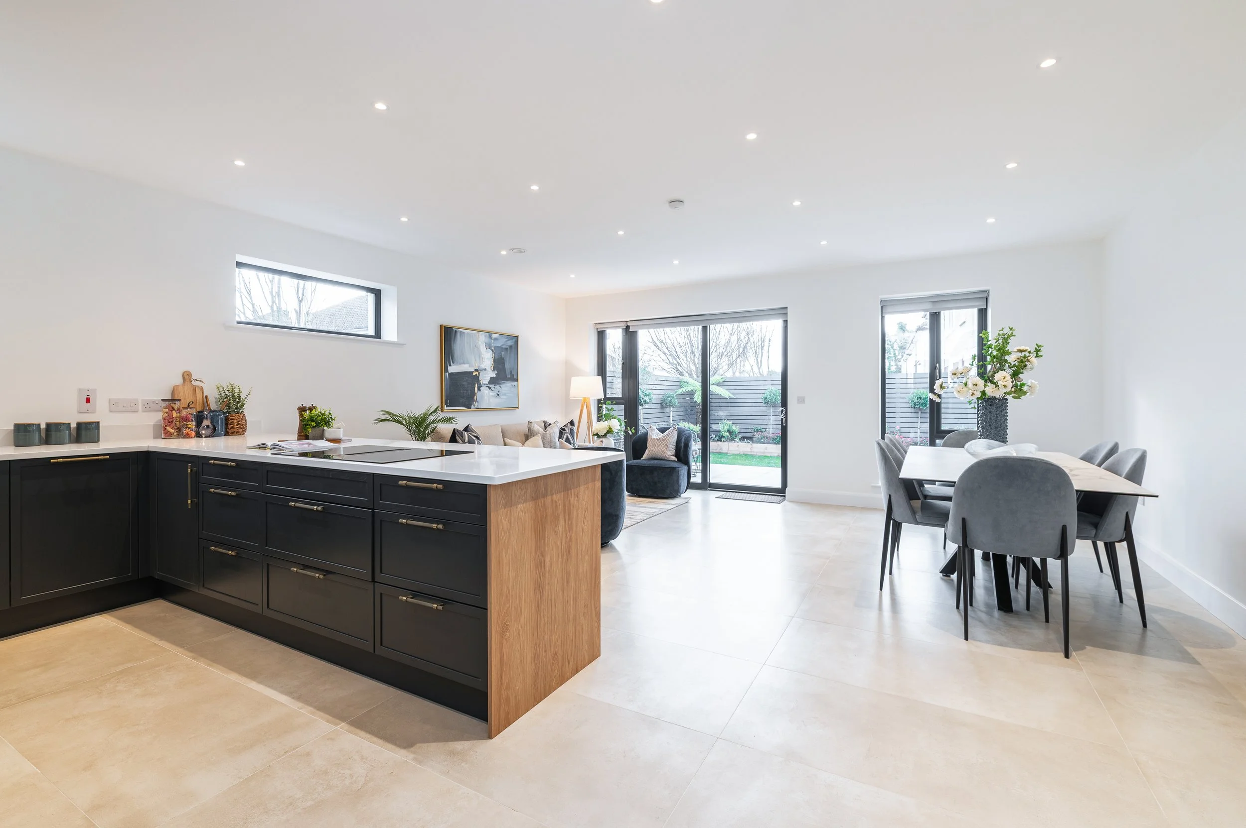 Open-concept living and dining area with black kitchen cabinets, a white countertop, and a table with gray chairs. Large windows and sliding doors lead to the backyard with some greenery. Decor includes a painting, flowers, and potted plants.
