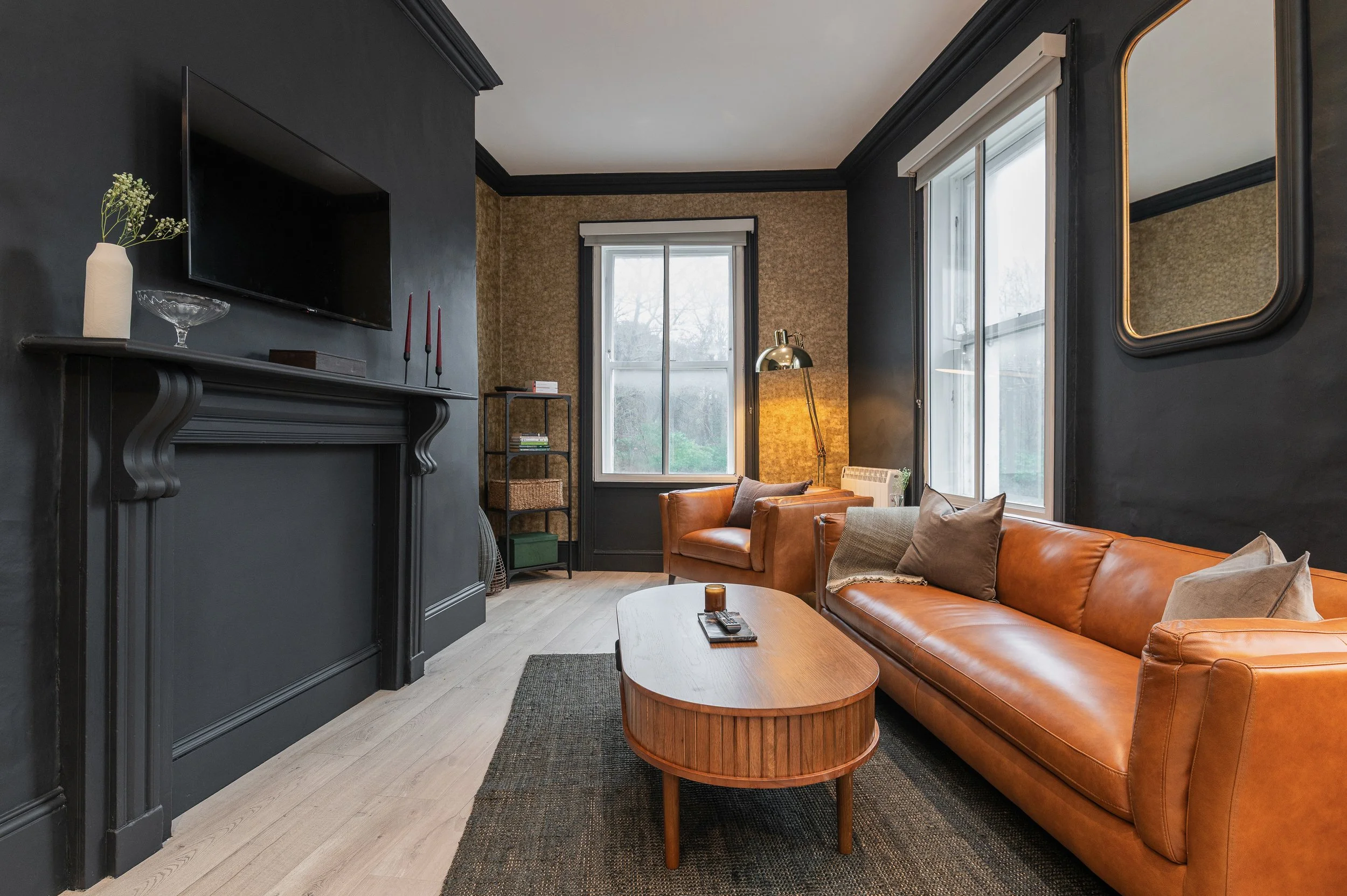 Living room with black accented walls, large windows, a leather sofa, a wooden coffee table, on which remotes and a candle sit, and a fireplace with a decorative vase and candles.