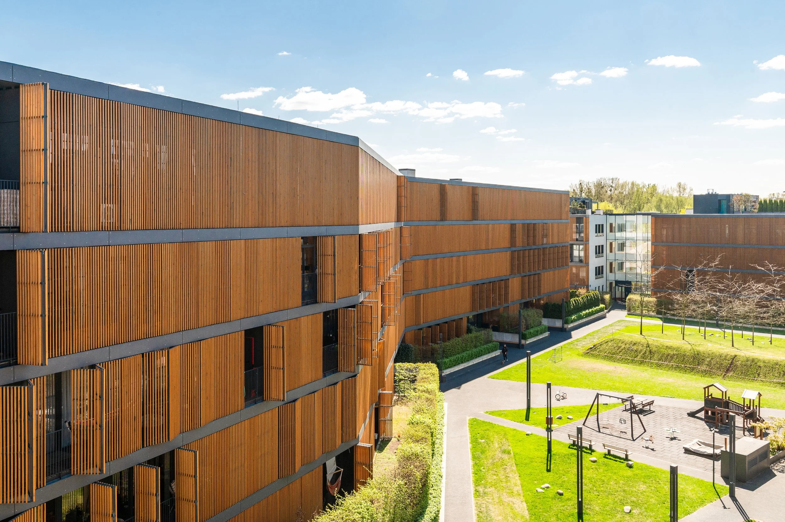 Modern building with wooden slat exterior and gray horizontal accents, with a green park area underneath and clear blue sky.