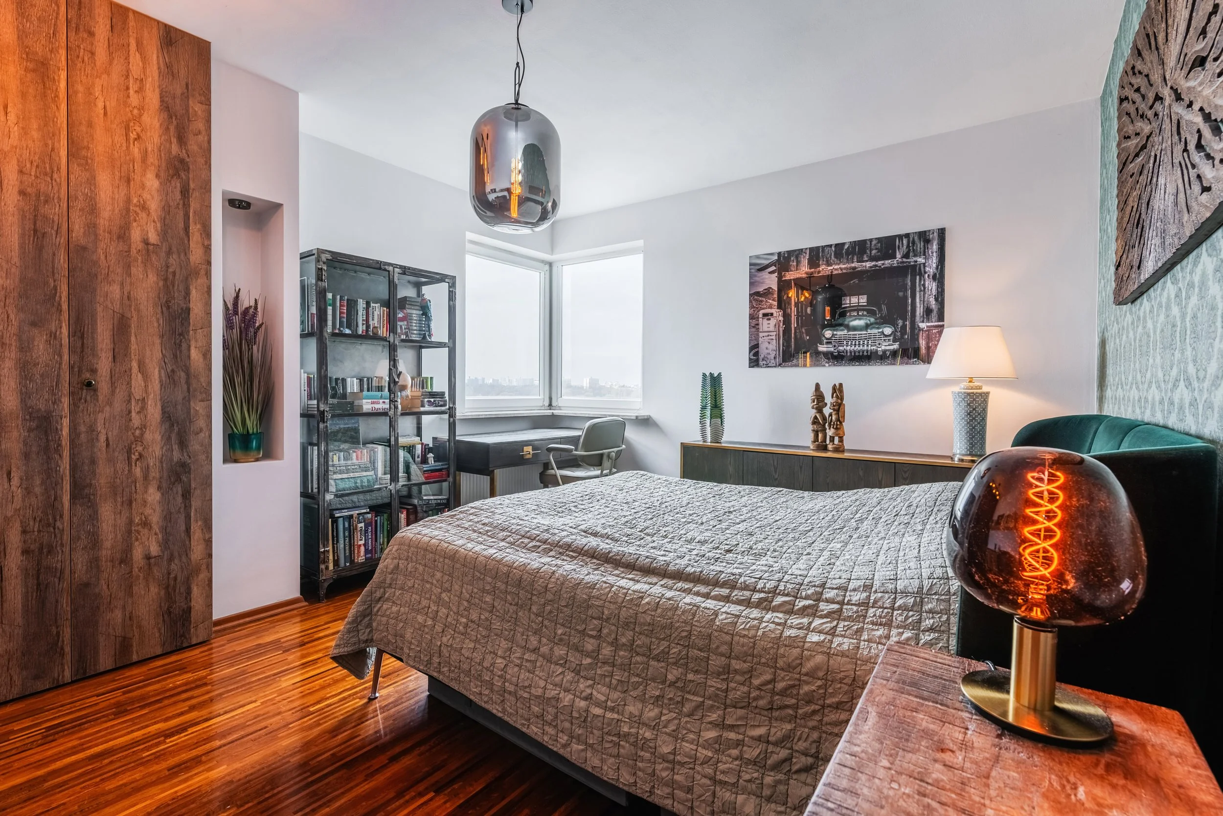 A modern bedroom with a bed covered in a beige quilt, a wooden wardrobe, a metal bookshelf filled with books, a desk with a chair, decorative wooden figurines and a plant on top, a large window with a city view, a black and white car wall art, a tabl