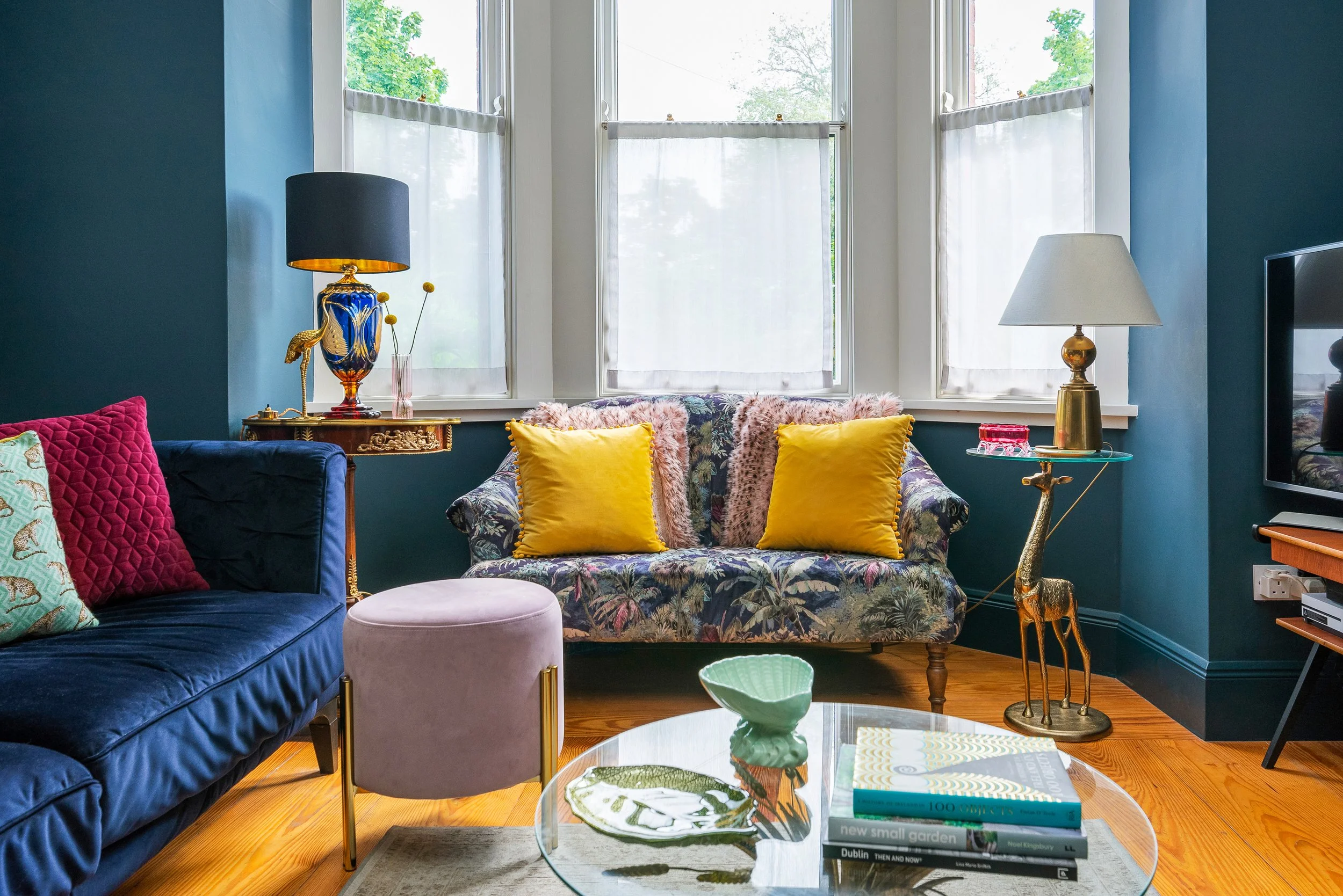 Living room with a floral sofa, yellow pillows, a glass coffee table with books and decorative items, and blue walls. Large windows with white curtains let in natural light.