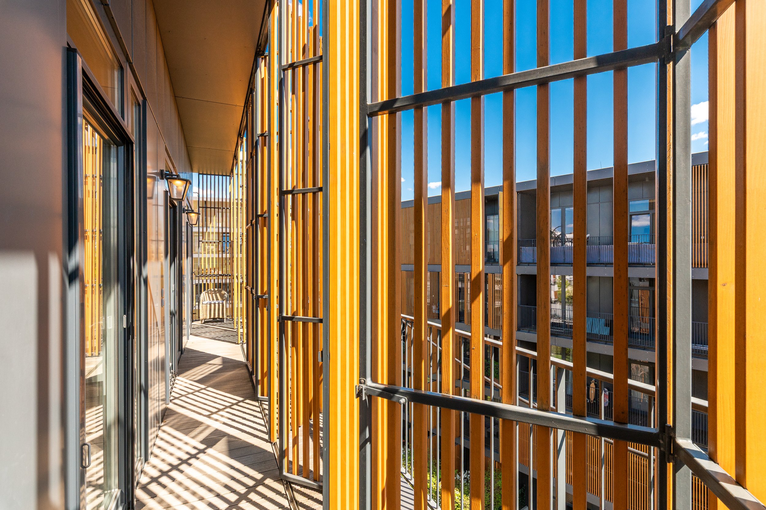 Balcony with orange and yellow vertical wooden slats, black metal railing, and glass door reflections, under a blue sky.
