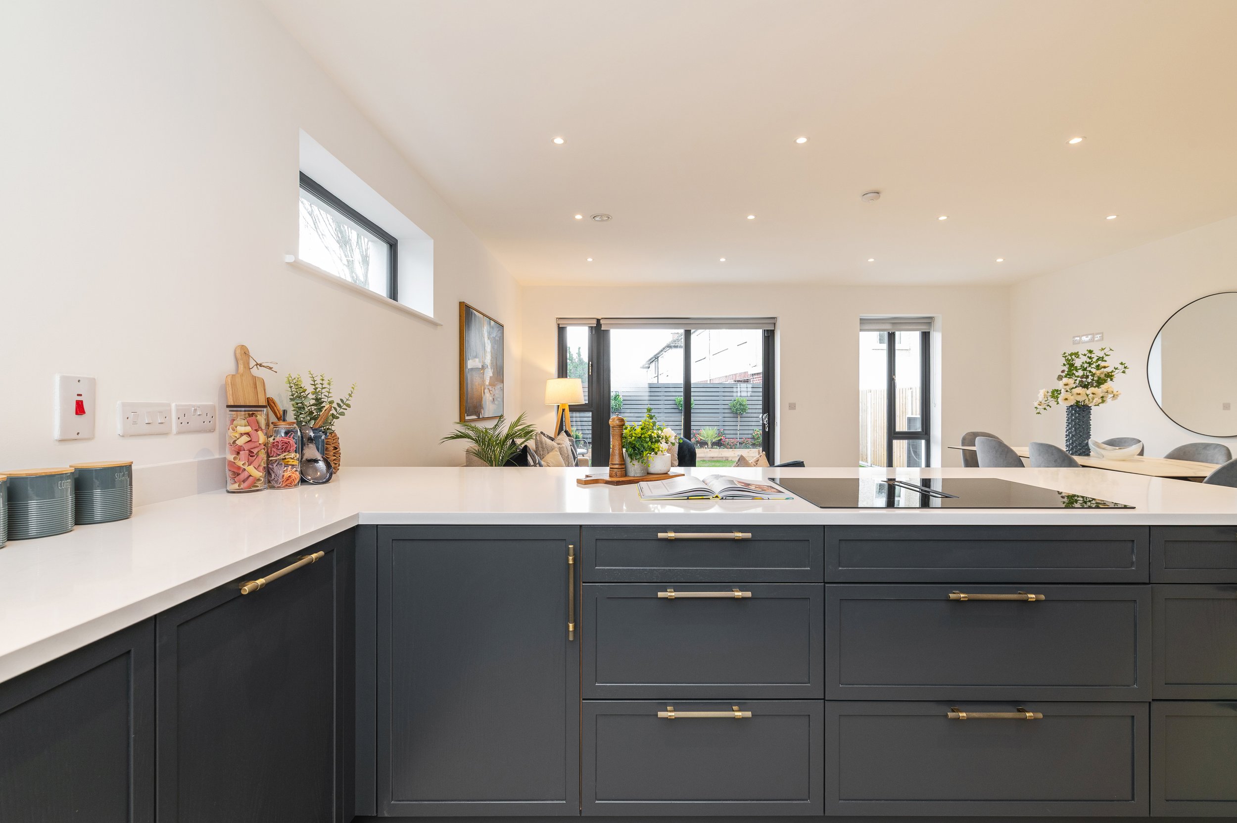 Modern kitchen with dark lower cabinets, white countertop, and various decorative items including jars and a potted plant. Bright, spacious dining and living area with large glass doors and plants visible outside.