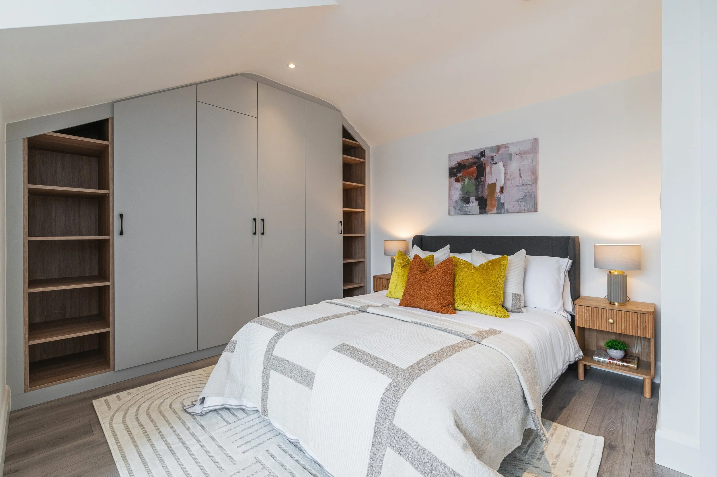 A modern bedroom with a bed that has a gray headboard, white bedding, and colorful pillows. On each side of the bed are wooden nightstands with lamps. There are white cabinets and open wooden shelves on the wall behind the bed, with a colorful abstra
