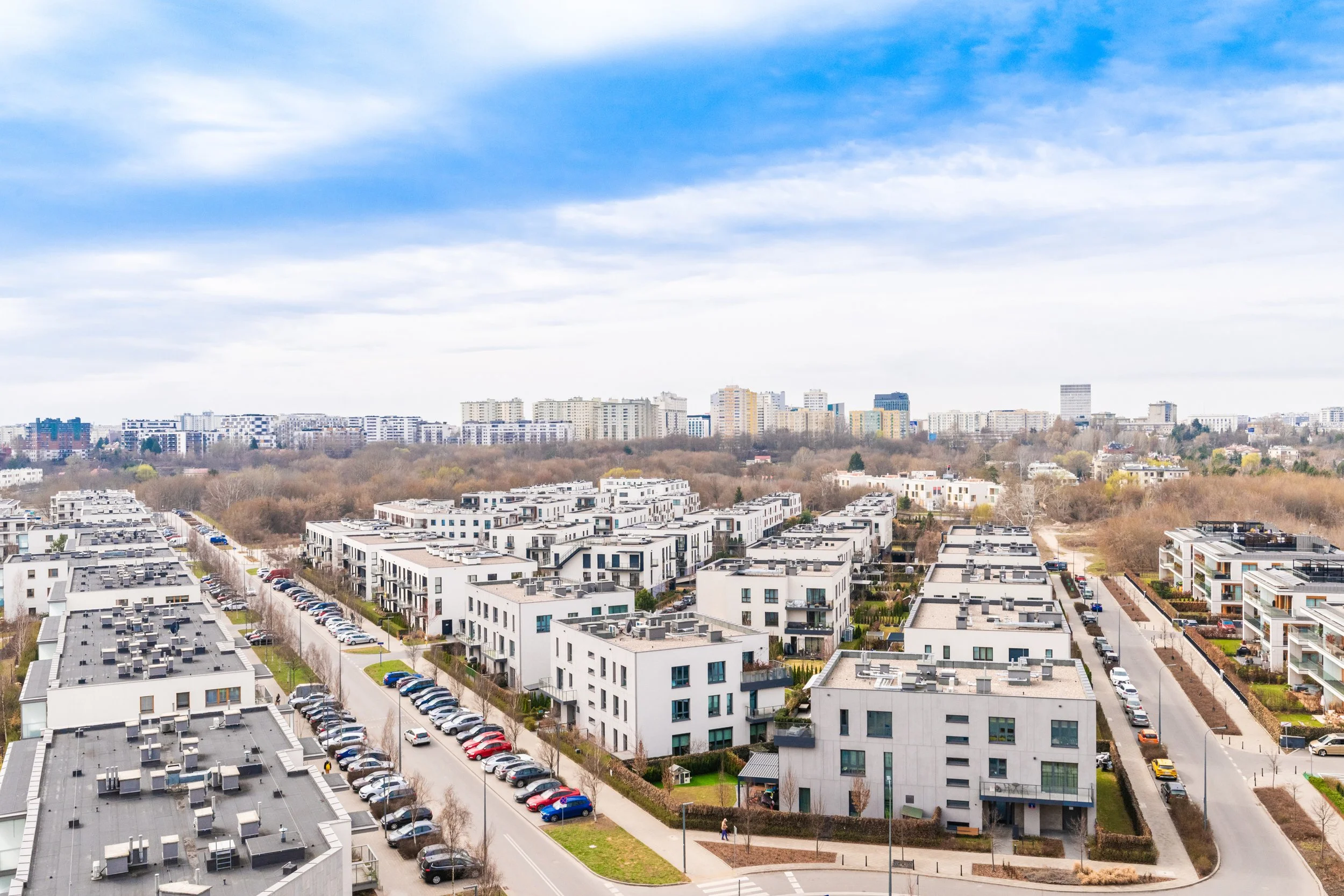 Urban cityscape with modern white apartment buildings, parking lot, and streets in the foreground, with a skyline of taller buildings and trees under a partly cloudy sky.
