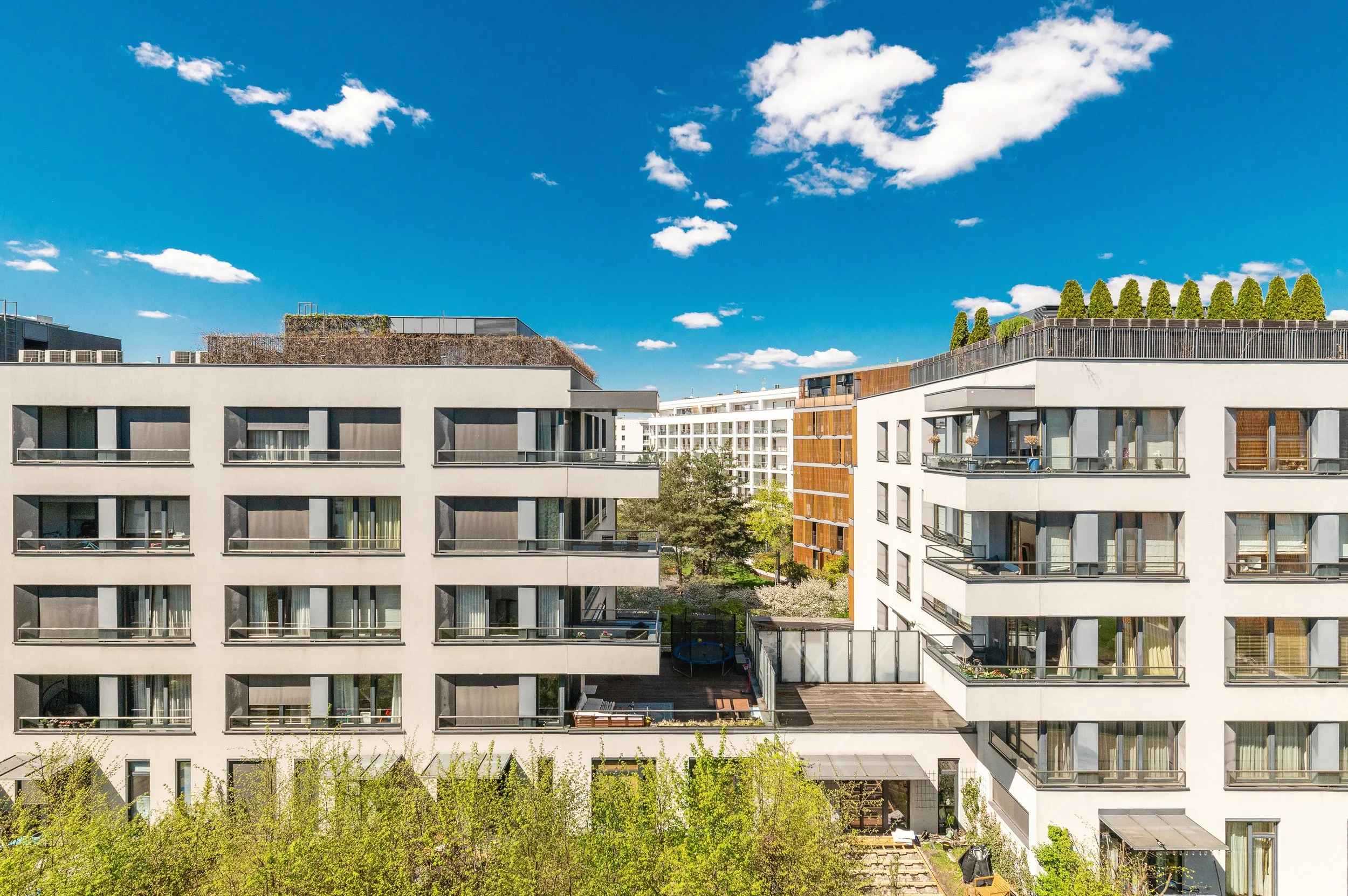 View of modern apartment buildings with balconies and rooftop greenery, under a blue sky with scattered clouds.
