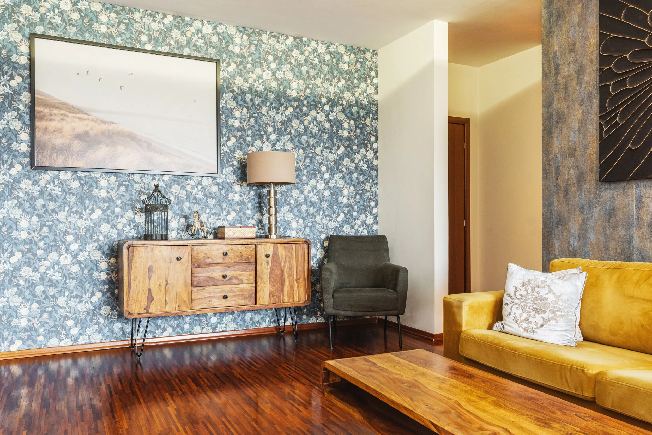 Living room corner with floral wallpaper, wooden dresser, gray armchair, yellow velvet sofa, artwork, lamp, and decorative items.