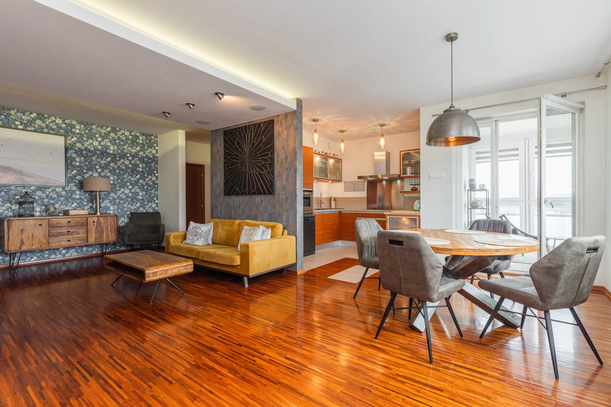 Open-concept living room and kitchen with hardwood floors, floral wallpaper accent wall, yellow sofa, round dining table with gray chairs, and large windows with balcony.