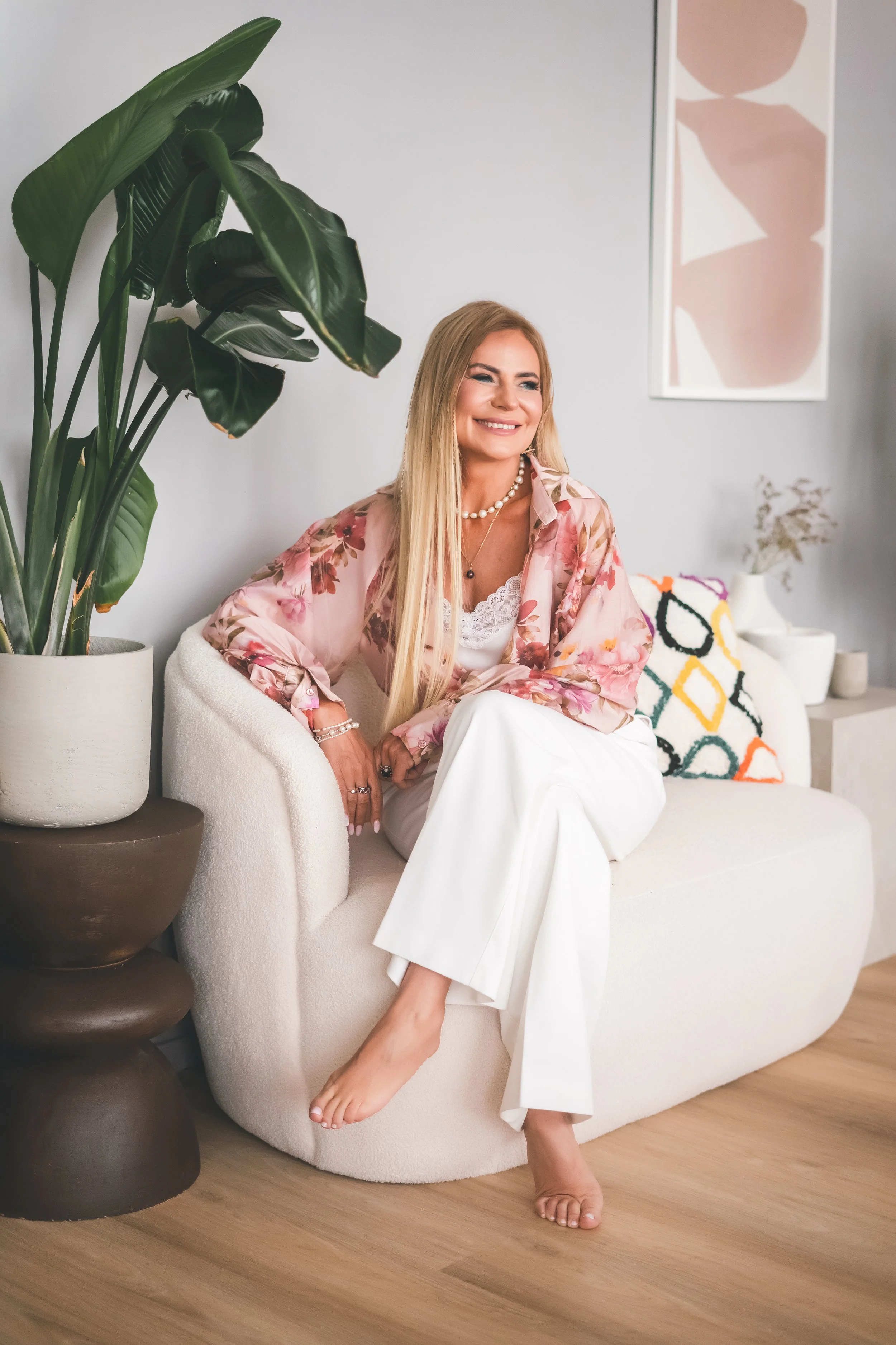 A smiling woman with long blonde hair sitting on a white, textured sofa. She is wearing a floral pink silk blouse, white dress pants, and jewelry including a pearl necklace, bracelets, and rings. Her right foot is resting on the sofa, and her left foot is on the floor, barefoot. To her left, there is a large potted plant with glossy green leaves. The background features modern wall art with shades of pink, beige, and white, and a decorative pillow with a colorful geometric pattern.