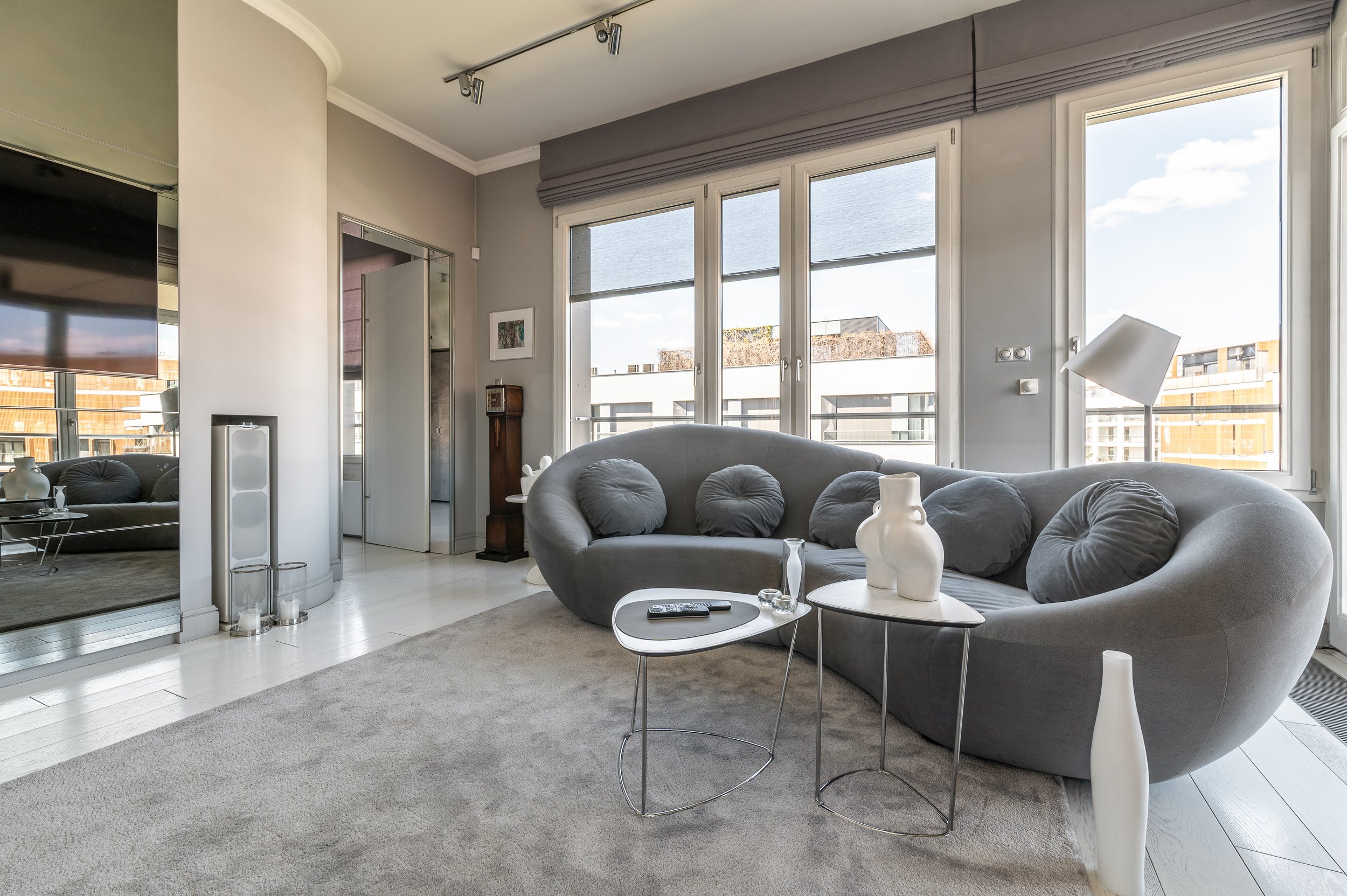 Modern living room with large windows, gray sofa with round pillows, white side tables with vases, and a mounted flat-screen TV