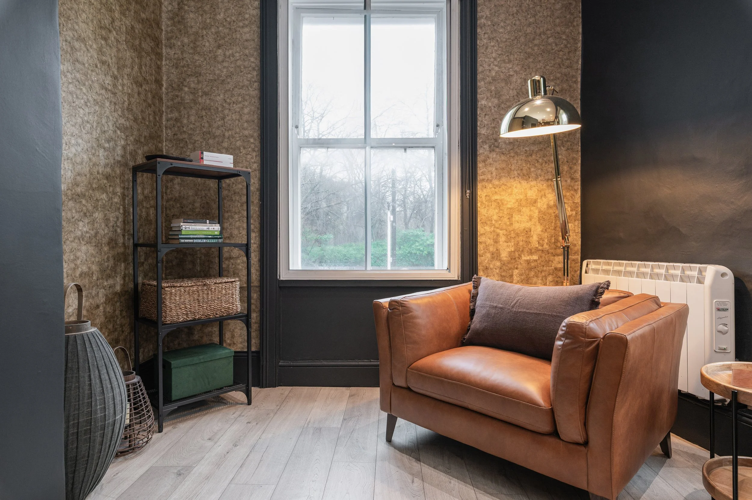 Living room with a large window, a brown leather sofa, a black metal bookshelf with books and woven basket, floor lamp with metallic shade, and a textured dark-colored wall.