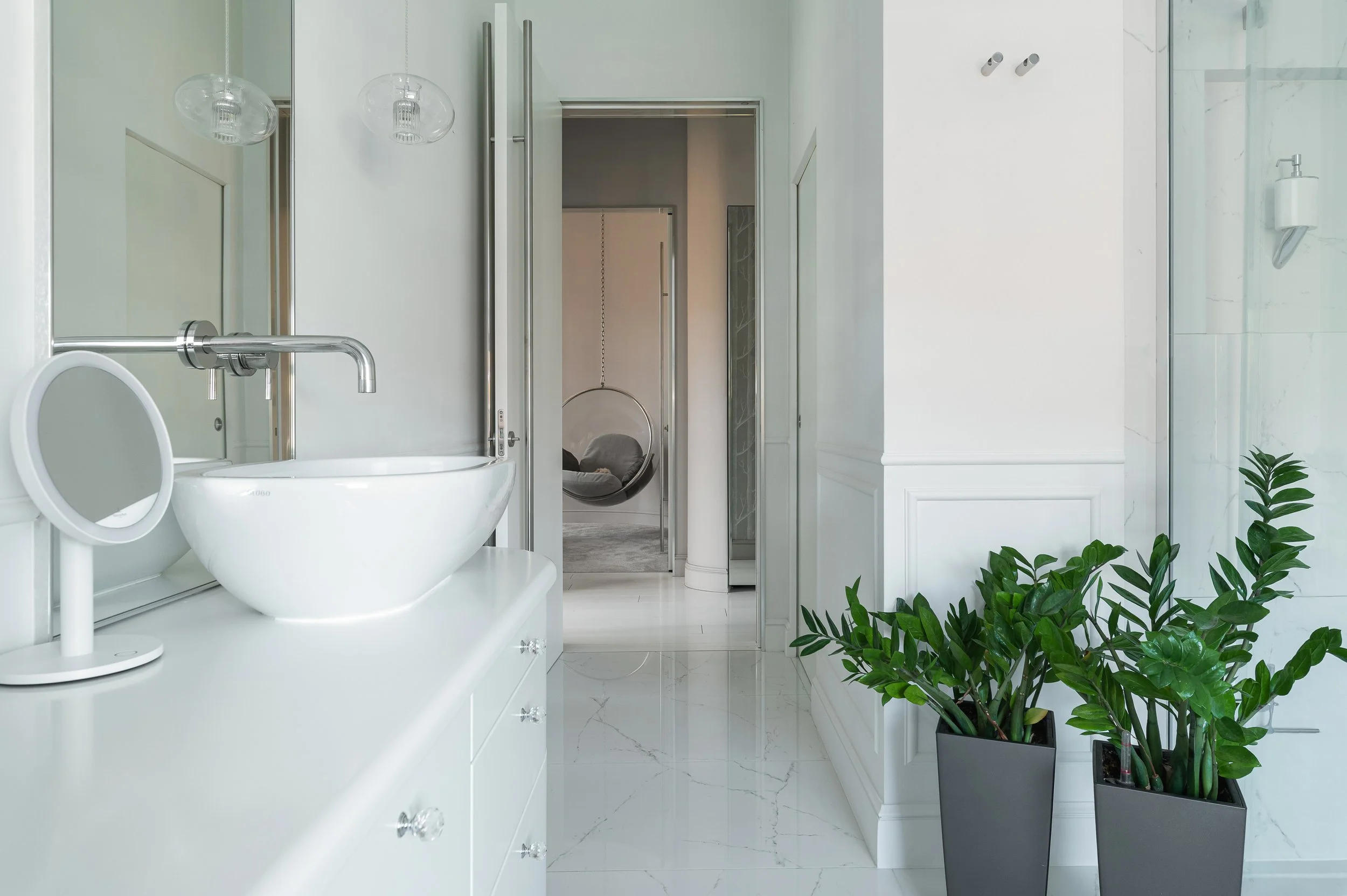 A modern bathroom with white cabinets, a vessel sink, a chrome faucet, decorative hanging light fixtures, potted green plants, and a doorway leading to a room with a hanging egg chair.