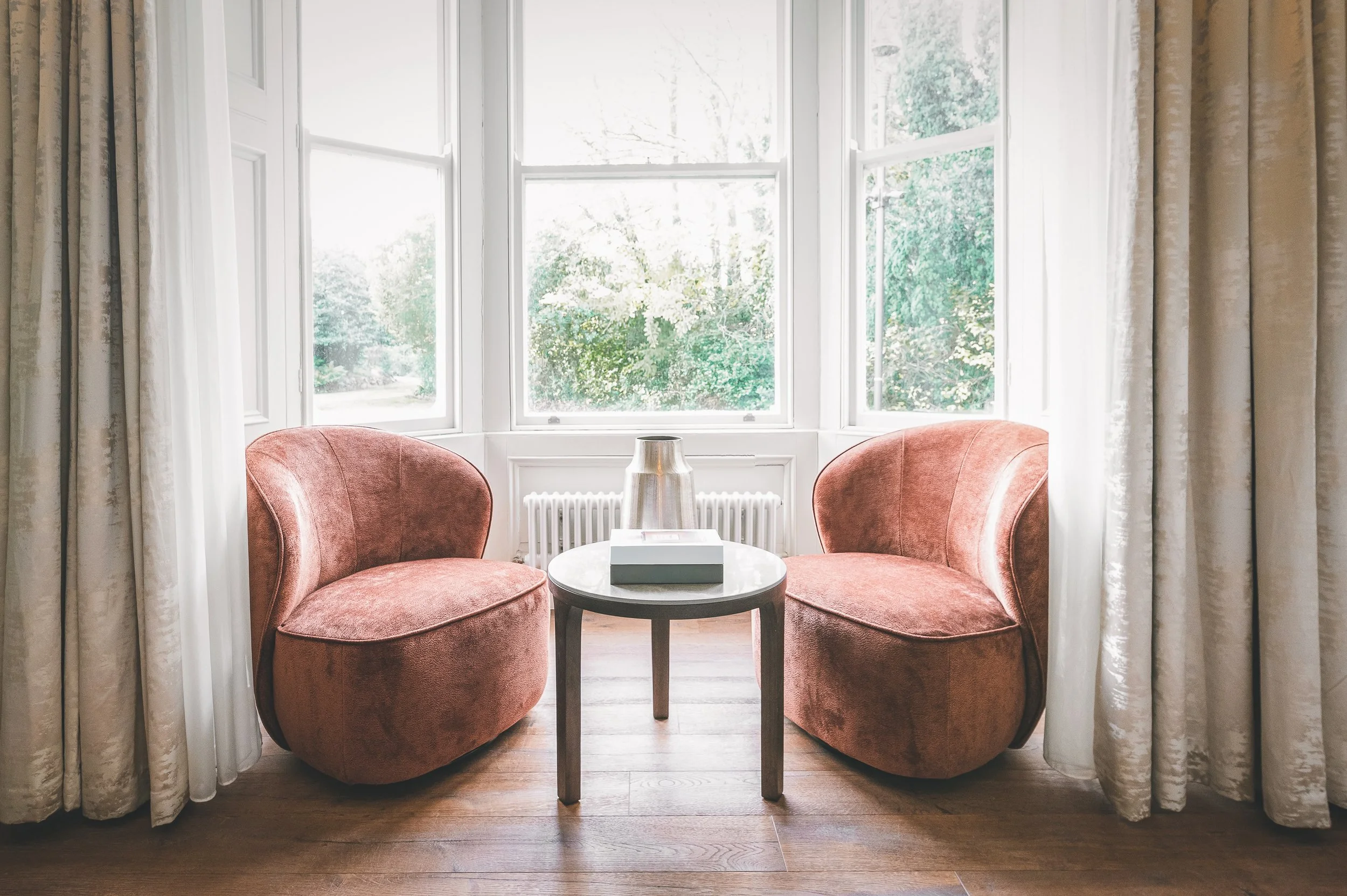 Two pink velvet chairs are positioned around a small dark wood round table with a box on top, situated in front of a large bay window with white framing and beige curtains on either side, overlooking trees outside.