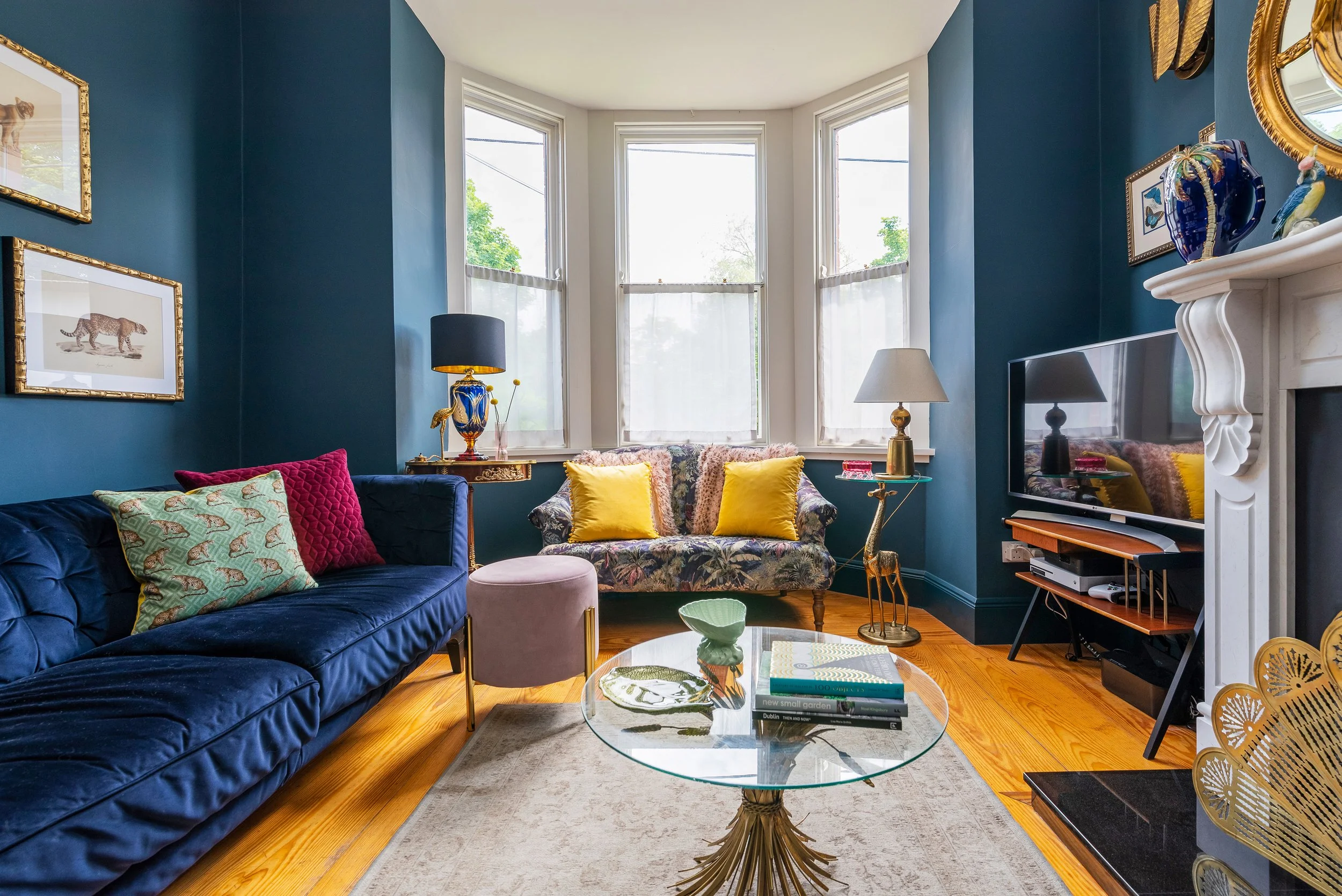 A living room with blue and beige walls, decorated with framed animal illustrations, gold accents, and colorful pillows. There is a velvet sofa, a patterned armchair with yellow pillows, a glass coffee table, and various decorative items including la