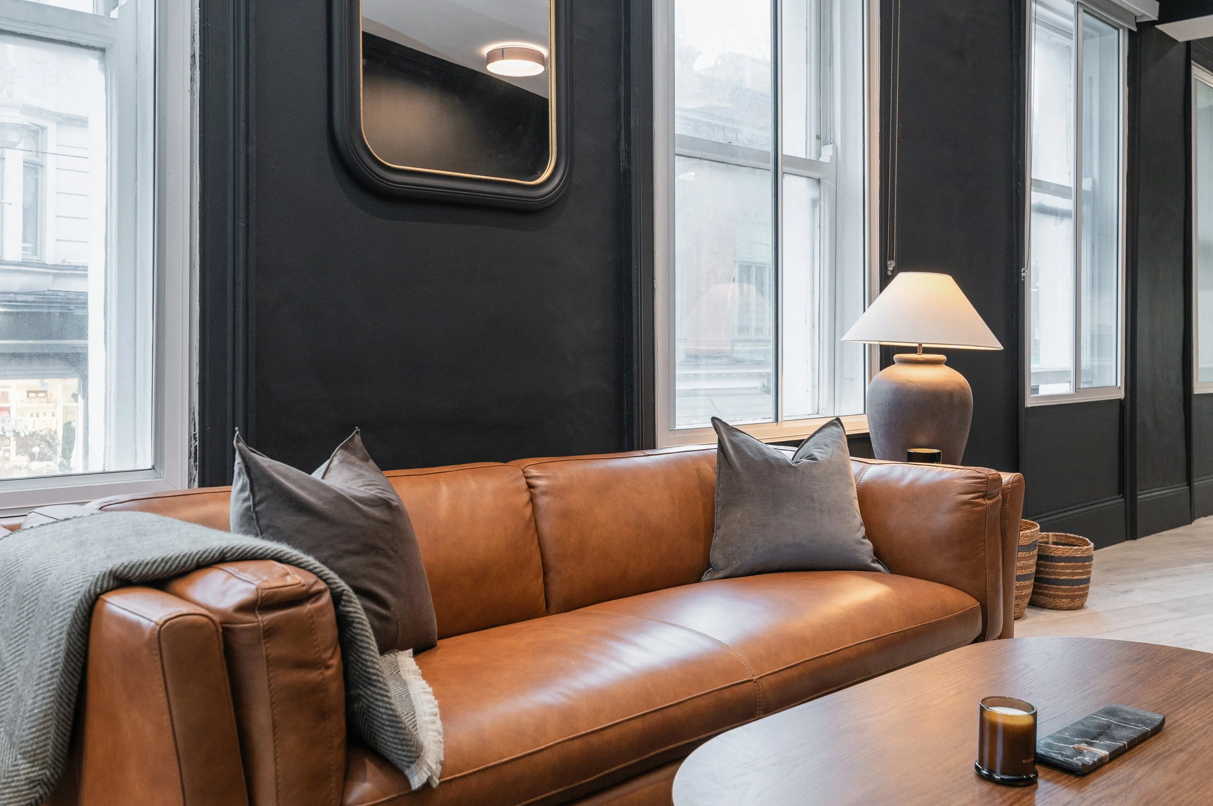 Modern living room with black walls, large windows, a leather sofa with pillows, a table with a candle, and a lamp.