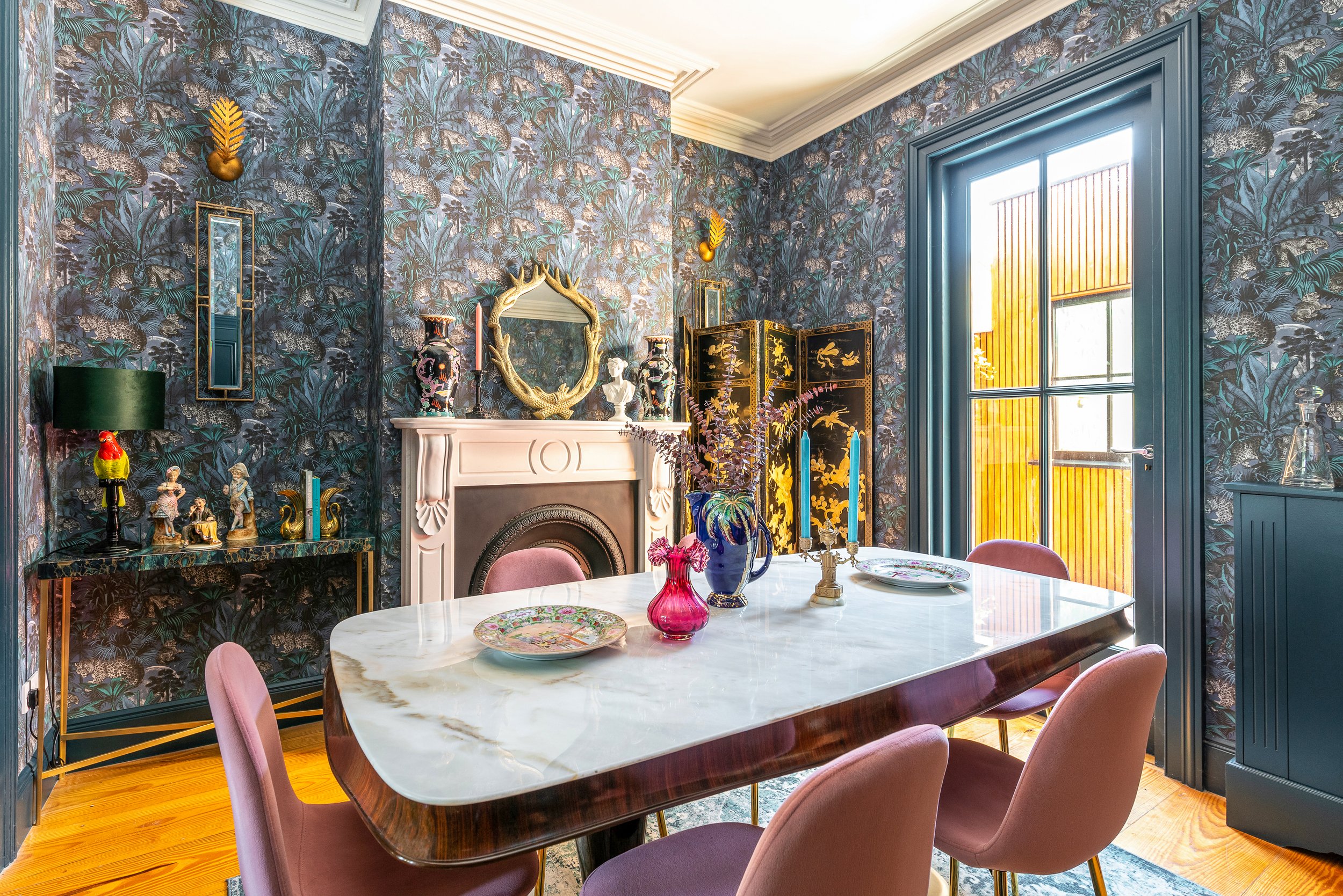 A dining room with dark tropical-themed wallpaper, a white fireplace mantel decorated with vases and sculptures, a gold-framed mirror, and a folding screen with black and gold design. The room has a large window with blue trim, pink upholstered chair