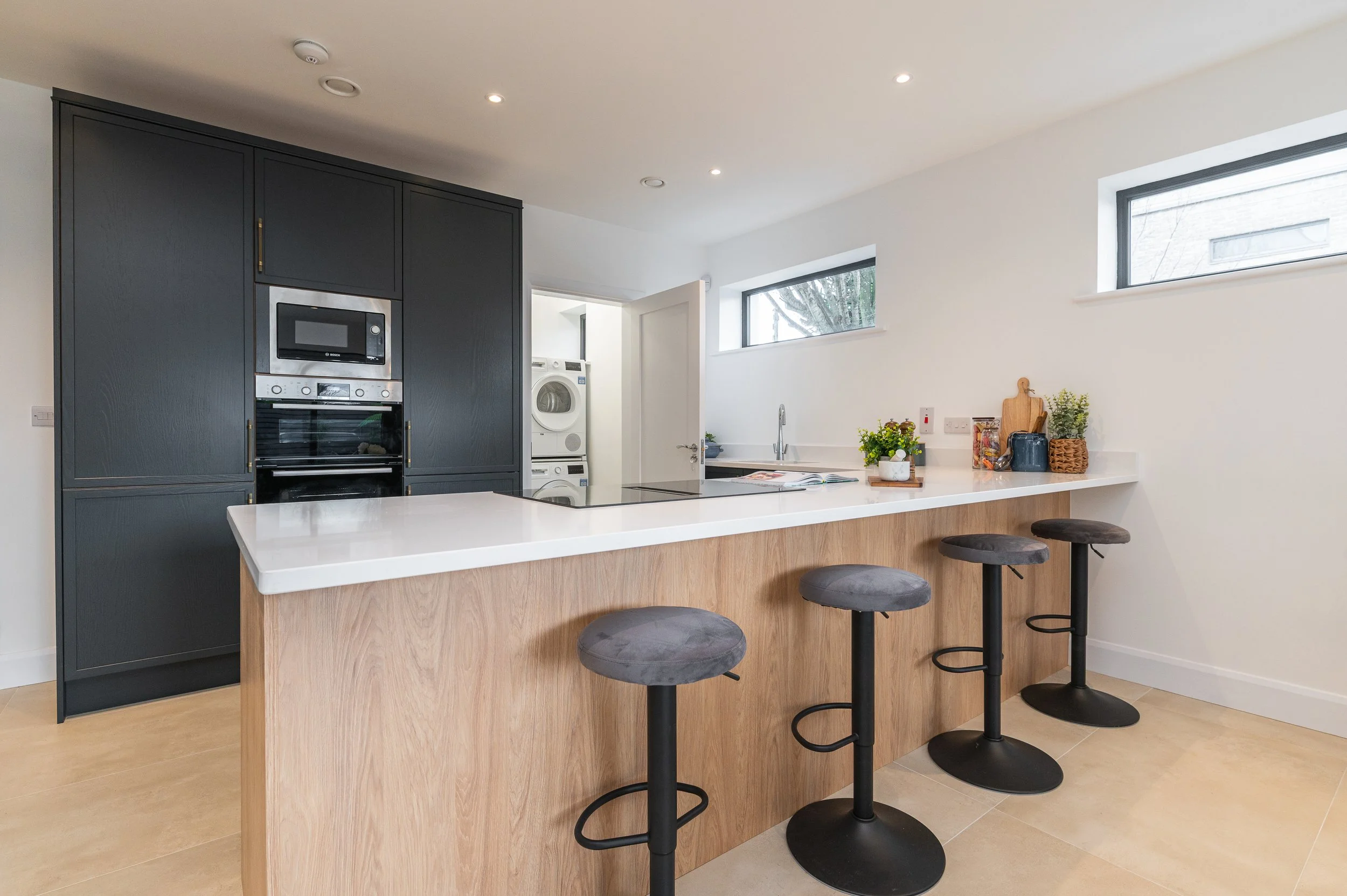 Modern kitchen with white countertop island, black bar stools, built-in oven and microwave, laundry area with washer and dryer, and two horizontal windows