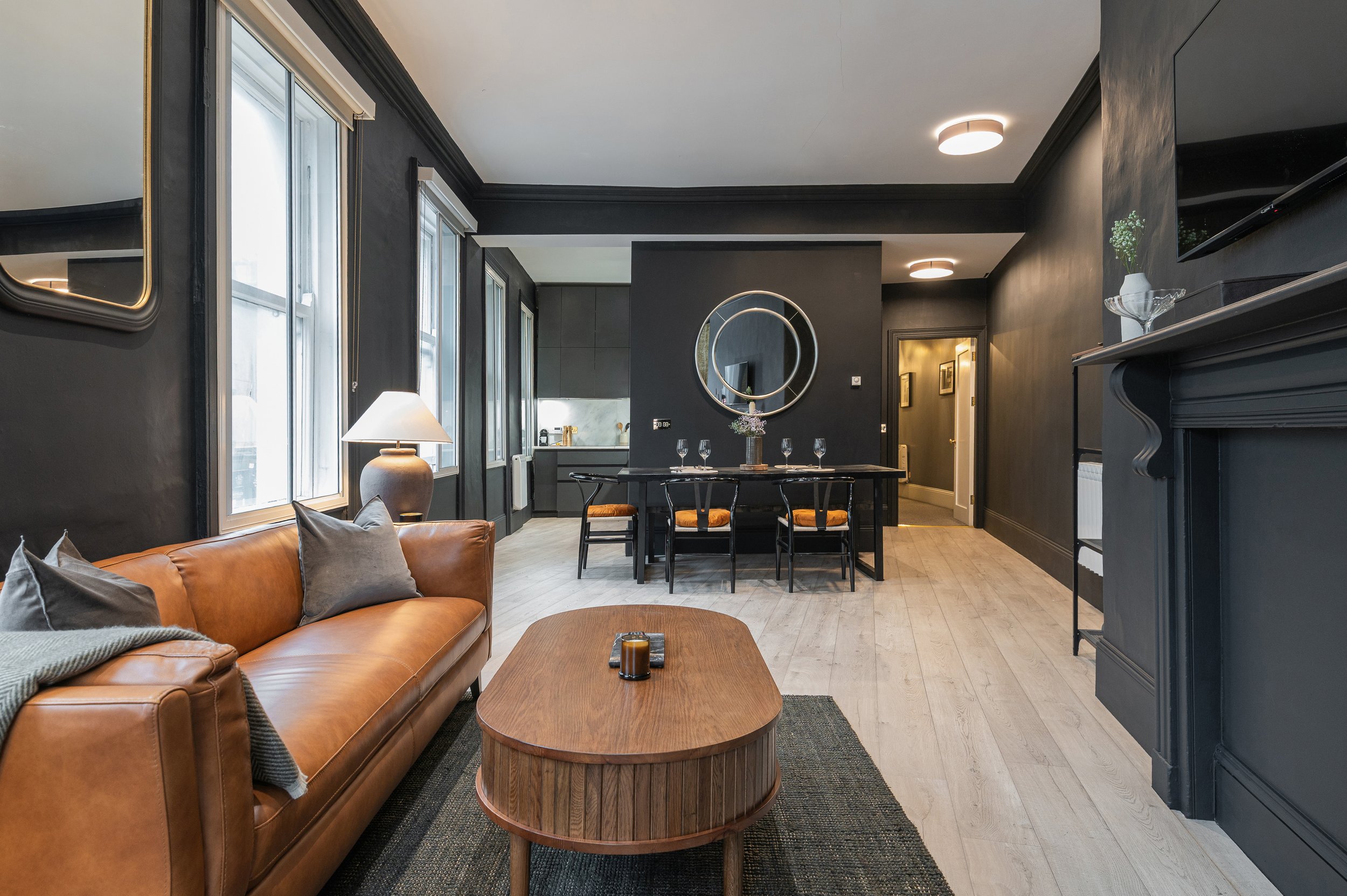 Modern living room with dark gray walls, large windows, a leather sofa with pillows, a wooden coffee table, and a dining area with a black table and four chairs, highlighted by ceiling lights and decorative mirrors.