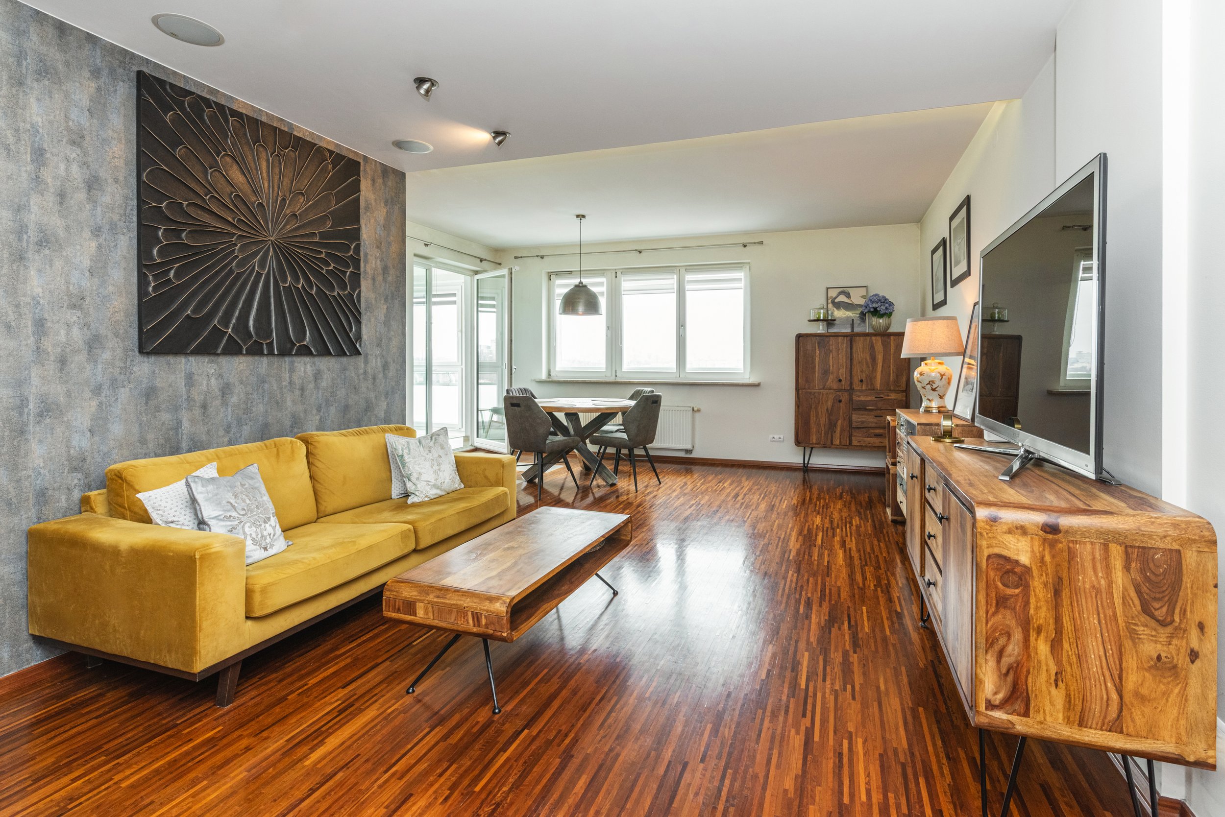 Living room with a yellow sofa, wooden coffee table, dining area with four chairs, window, and wall decorations.