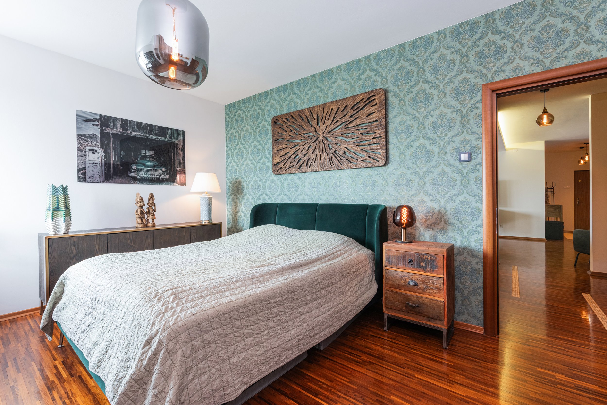 A bedroom with a bed, wooden side table, and decor on the wall. There is a doorway leading to another room with pendant lighting.