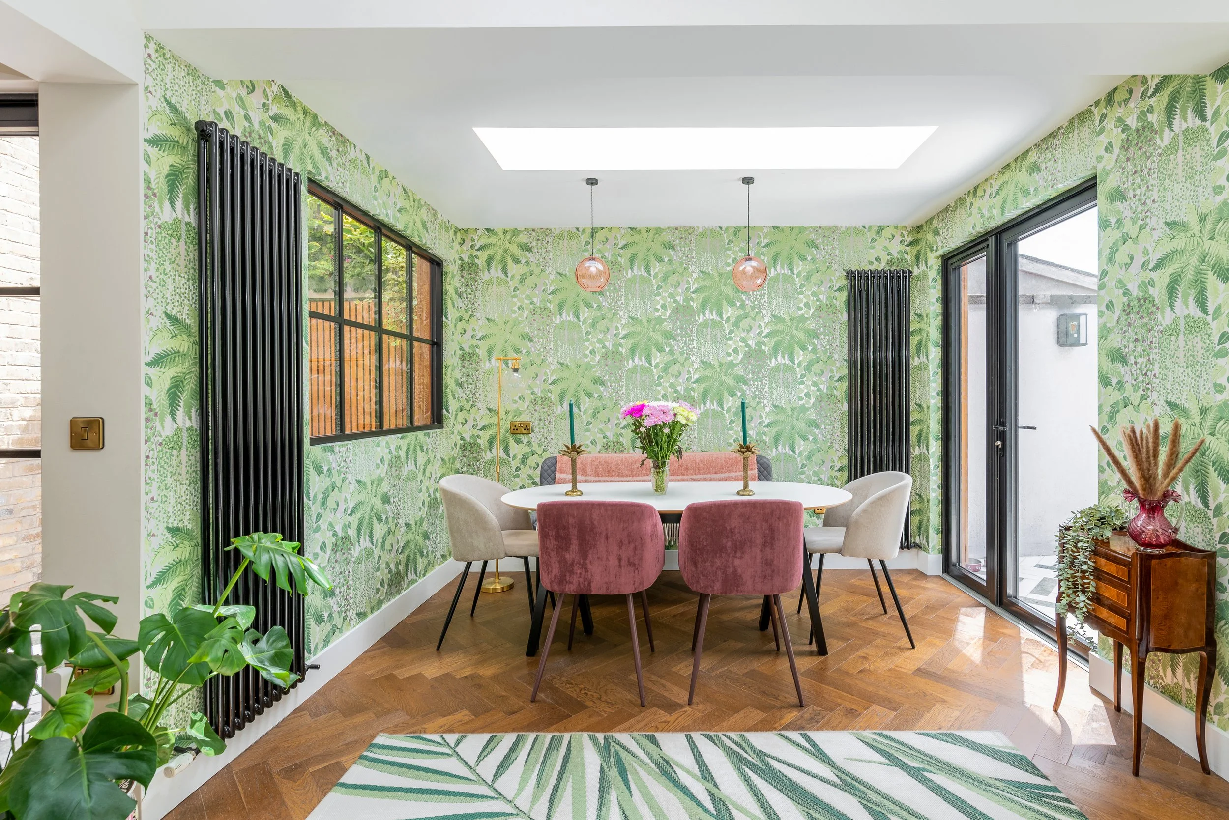 Dining area with tropical leaf patterned wallpaper, round white and pink velvet chairs, a white oval table with gold candle holders and a vase of pink and purple flowers, large windows, green plants, and natural light from a skylight.