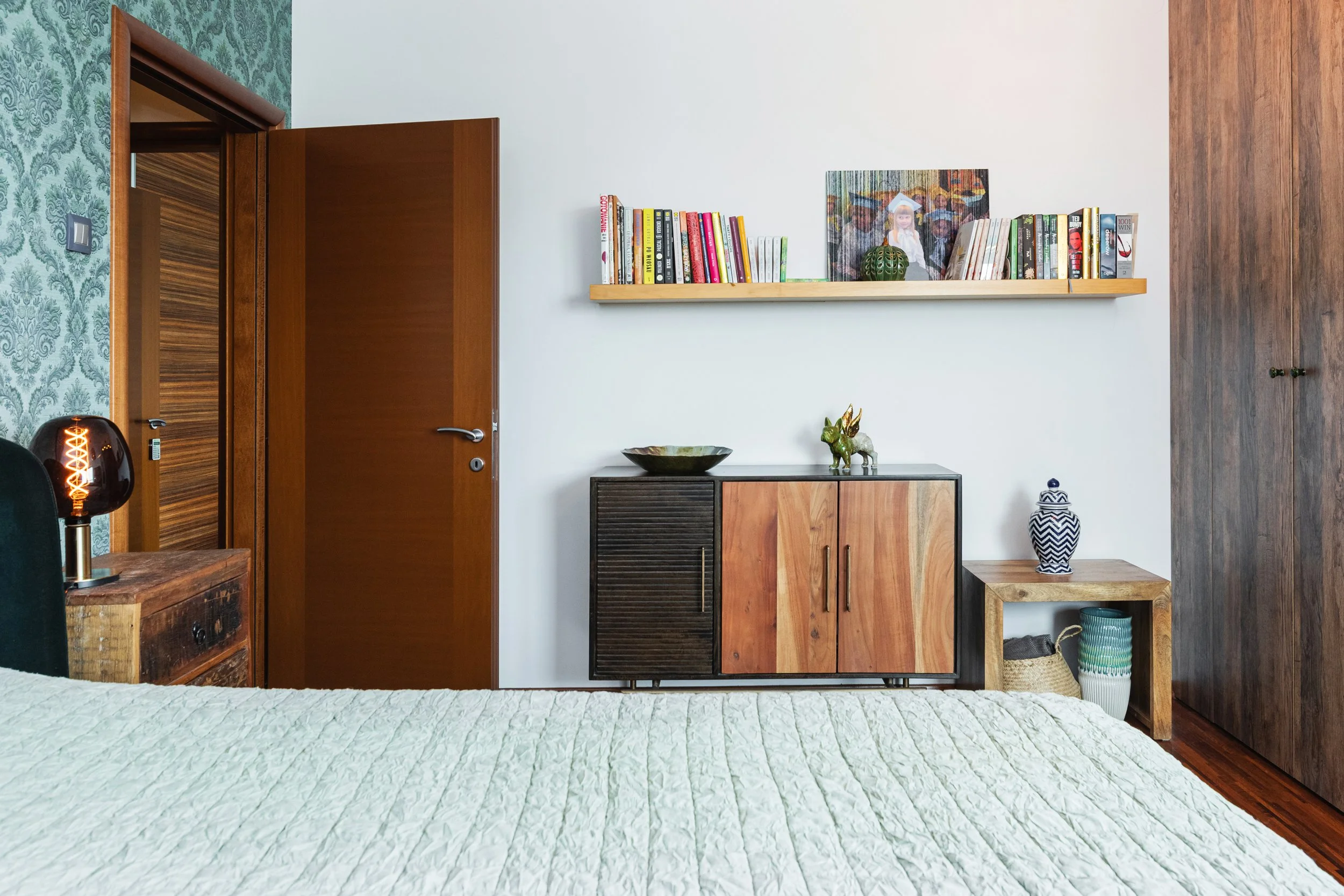 A room with a bed featuring a white quilt, a wooden side table with a dark lamp, a wall with patterned green wallpaper, a white wall with wooden shelves holding books and decorative items, a black and wood cabinet, and a vase with a blue and white de