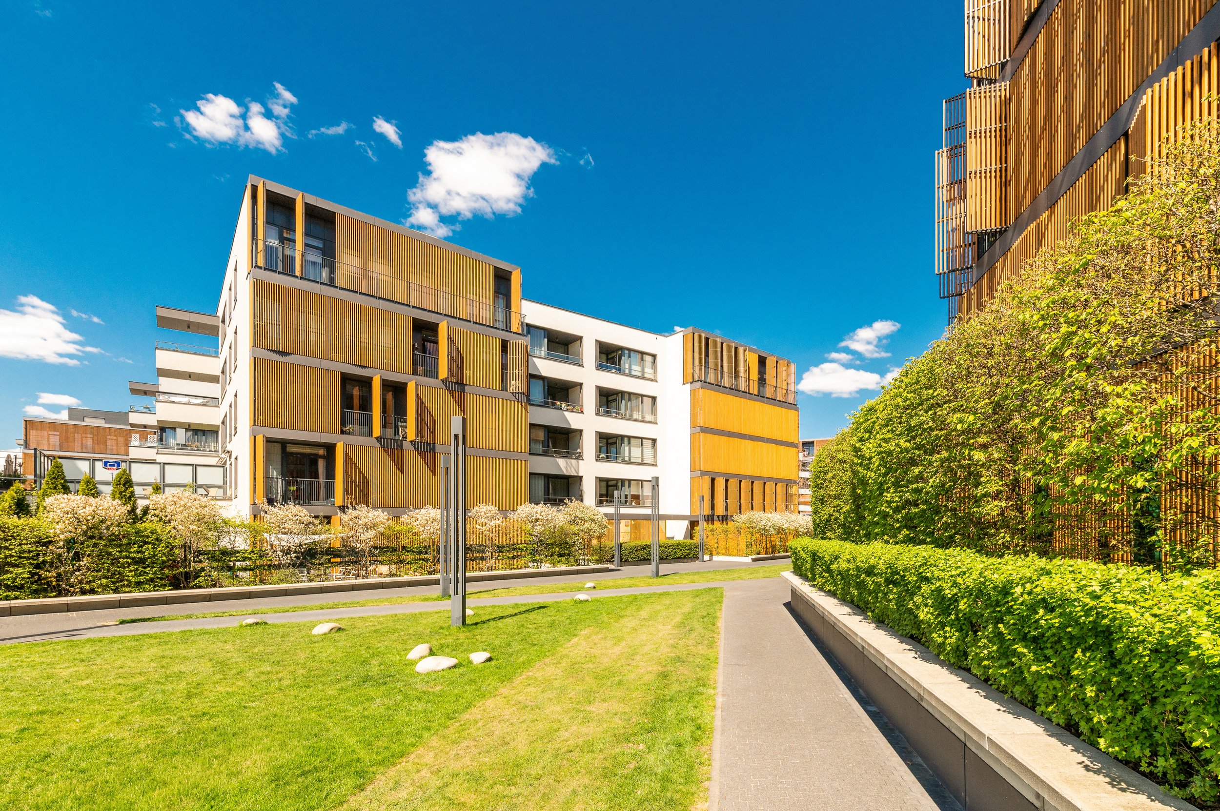 Modern multi-story apartment buildings with wooden slat accents, blue sky with white clouds, and landscaped green area with walkways and trees.