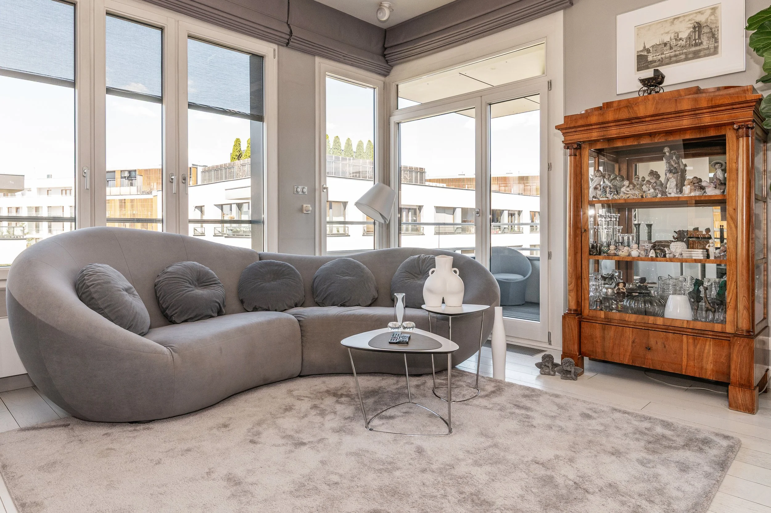 Living room with large windows, a gray curved sofa with dark gray cushions, a white vase on a small round table, a floor lamp, and a wooden display cabinet filled with porcelain figurines and glassware.
