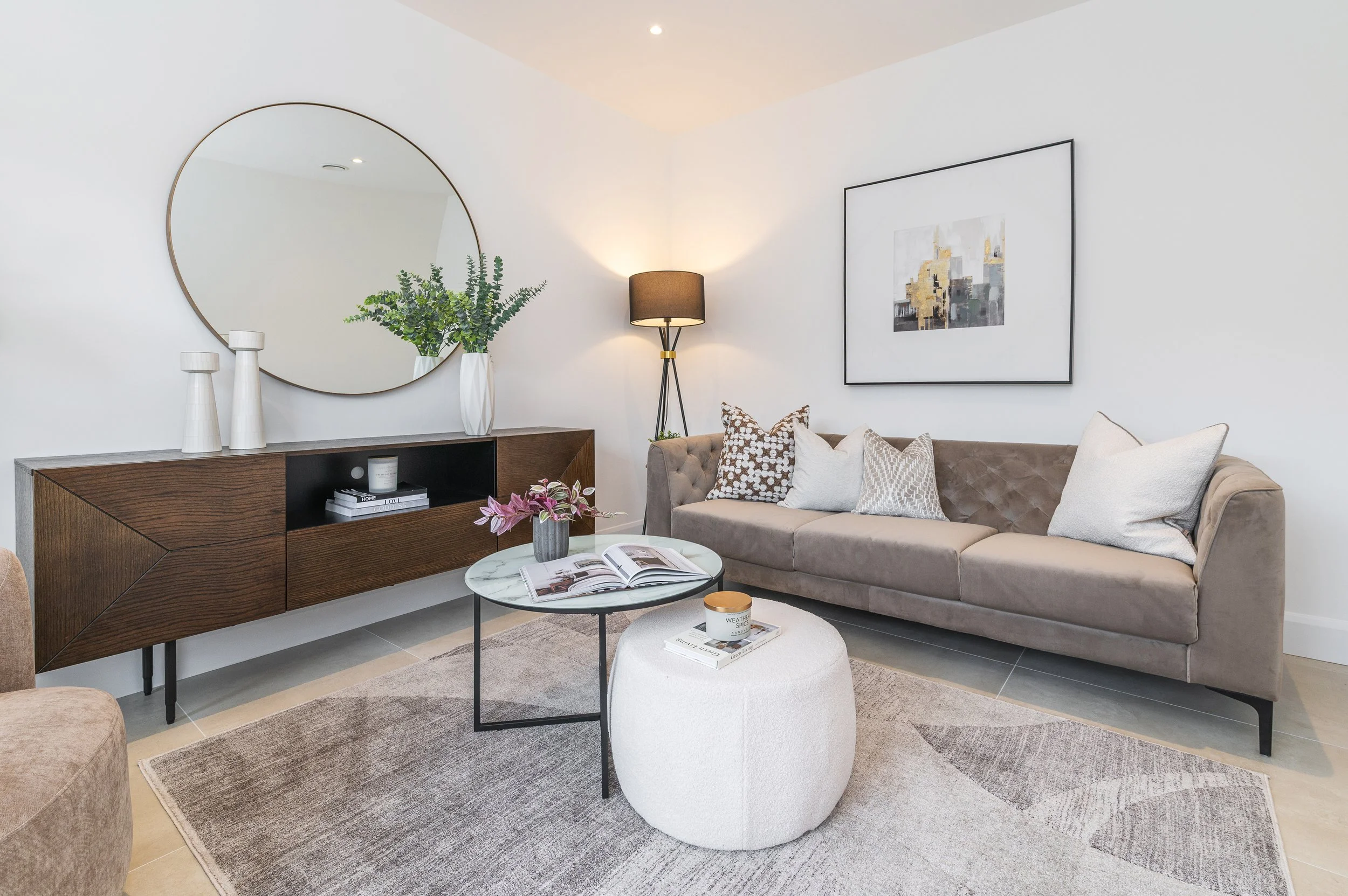 A modern living room with a beige tufted sofa decorated with patterned cushions, a round marble-topped coffee table with magazines, a white ottoman, a wooden sideboard with white vases and a plant, a large circular mirror, a framed abstract artwork, 