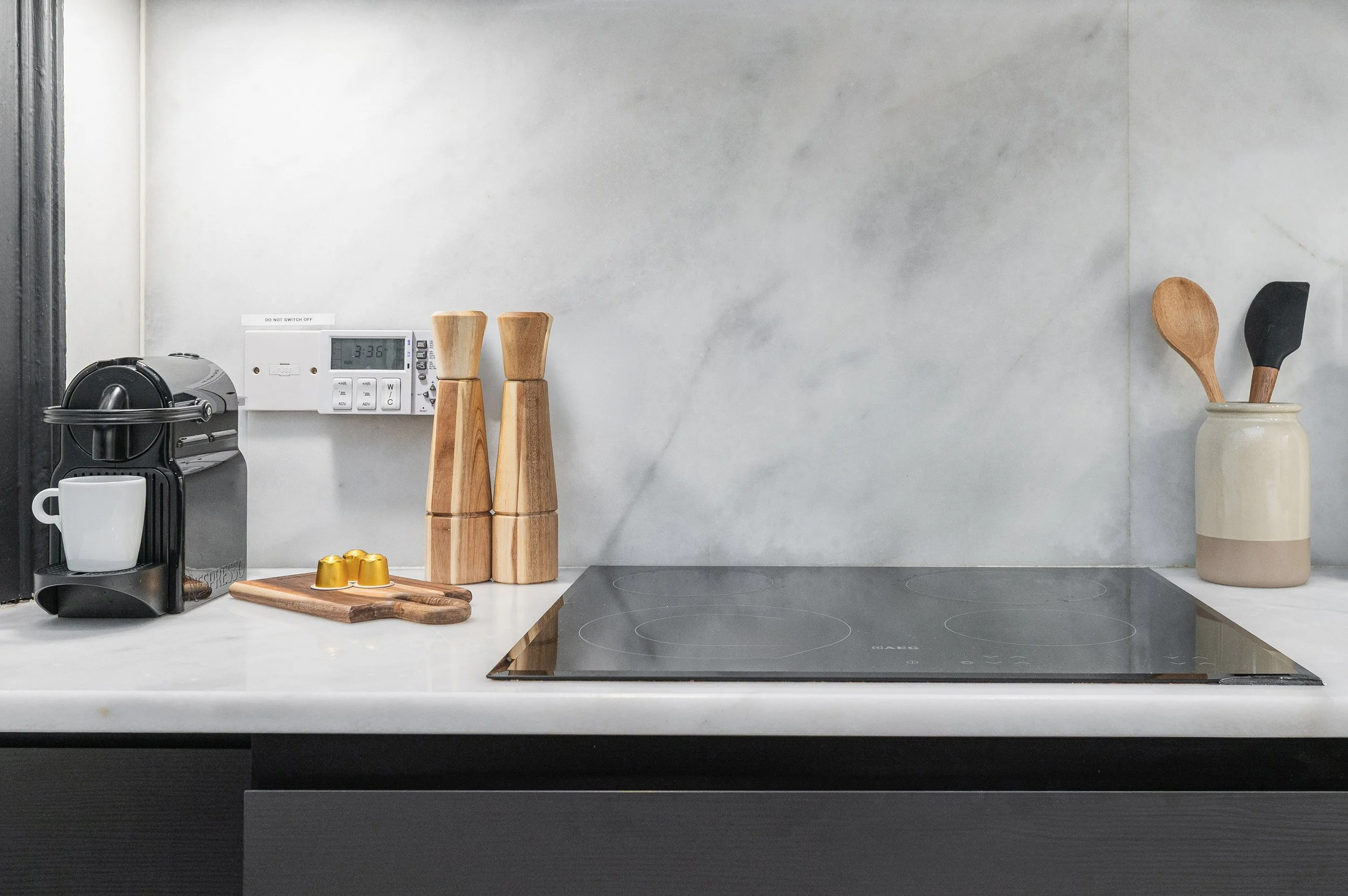 A kitchen countertop with a coffee machine, a white mug, wooden pepper and salt shakers, a small cutting board with golden capsules, a black countertop microwave, and a ceramic vase holding wooden and black spatulas. A white digital timer is mounted 