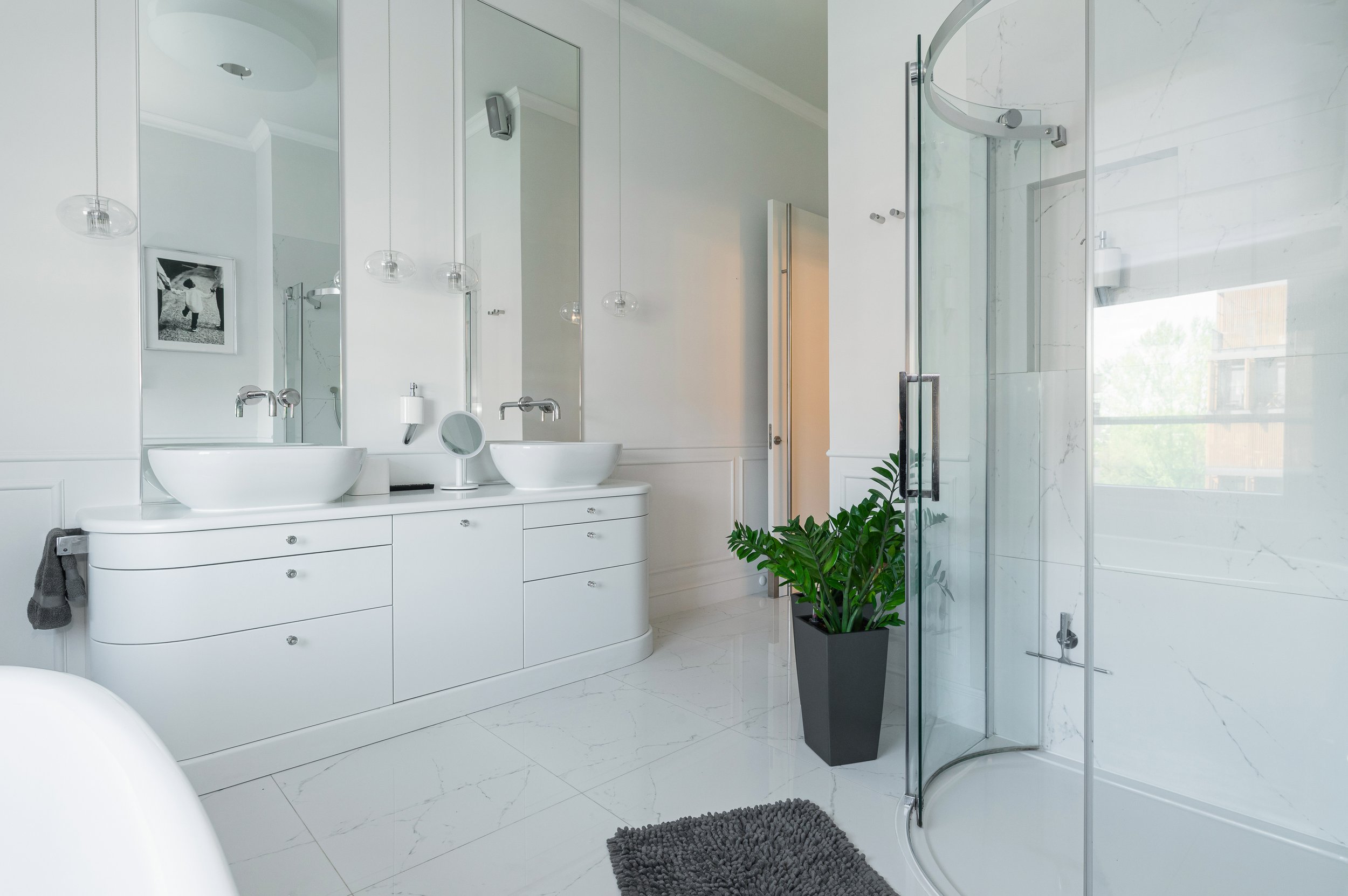Modern white bathroom with a large mirror, dual vessel sinks, a potted green plant, and a glass-enclosed shower with marble walls and a window.