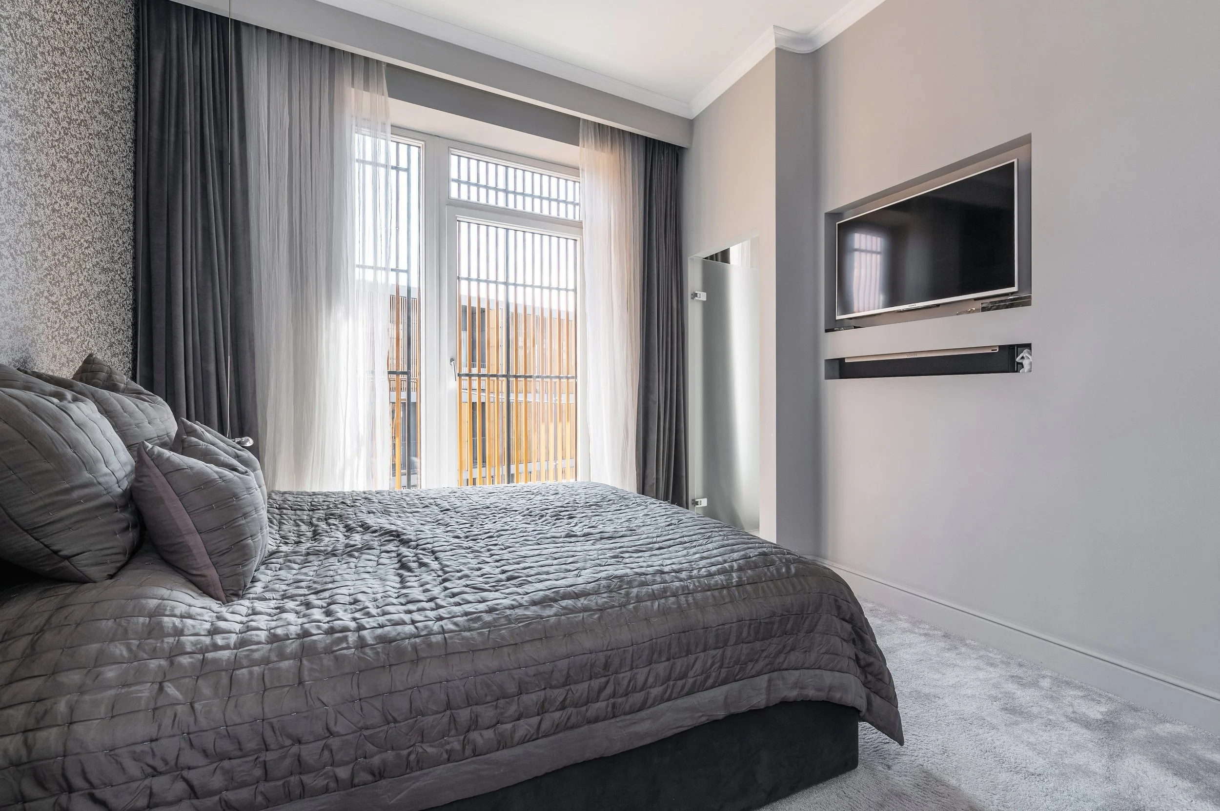 A modern bedroom with a large bed covered in a gray quilt and pillows, gray curtains, a window with vertical blinds, a flat-screen TV on the wall, and a carpeted floor.