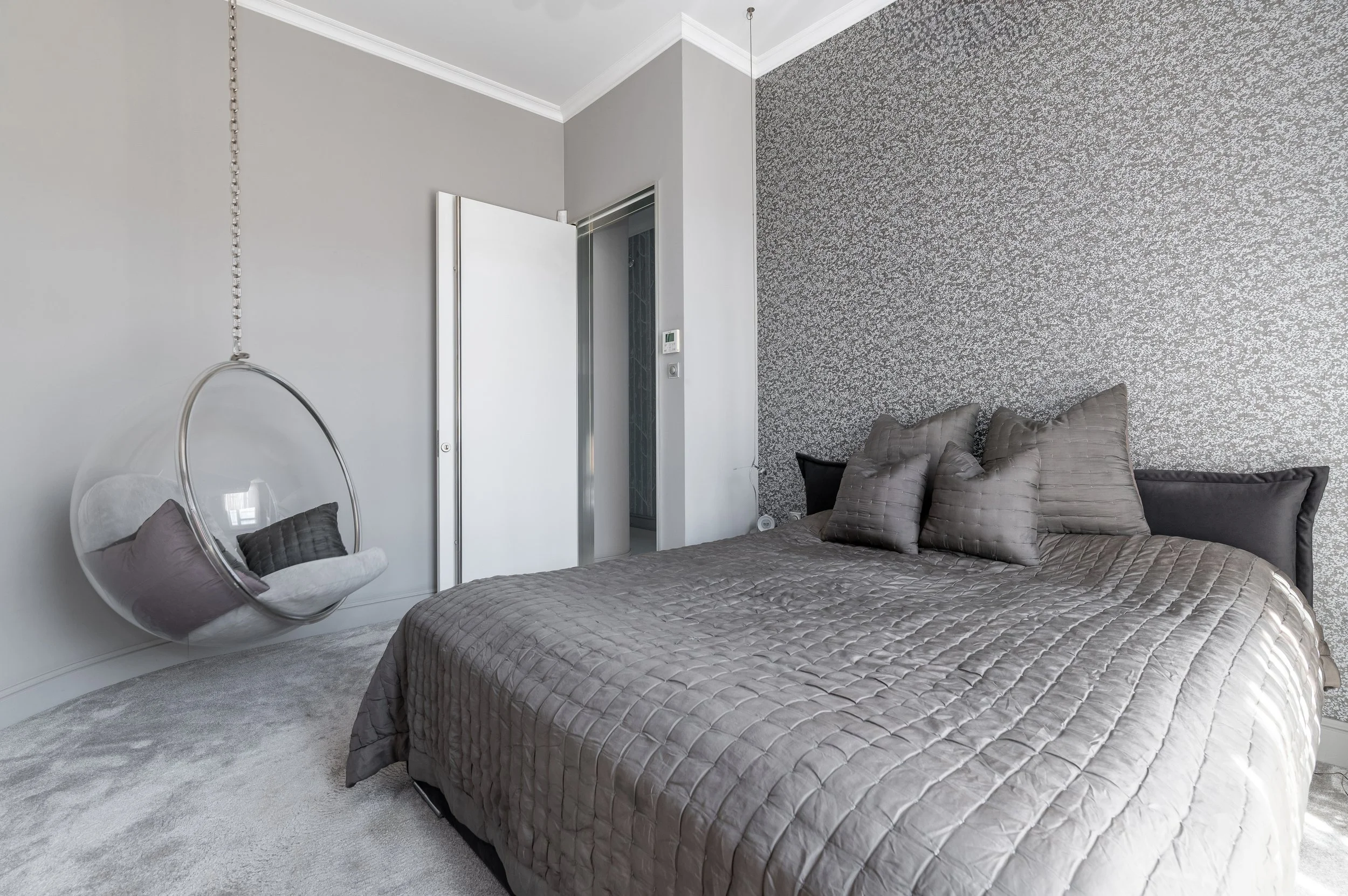 A bedroom with a large bed covered in a quilted taupe bedspread and matching pillows. To the left, there is a hanging transparent bubble chair with additional pillows inside. The room has a white ceiling, crown molding, and two-toned walls with one s