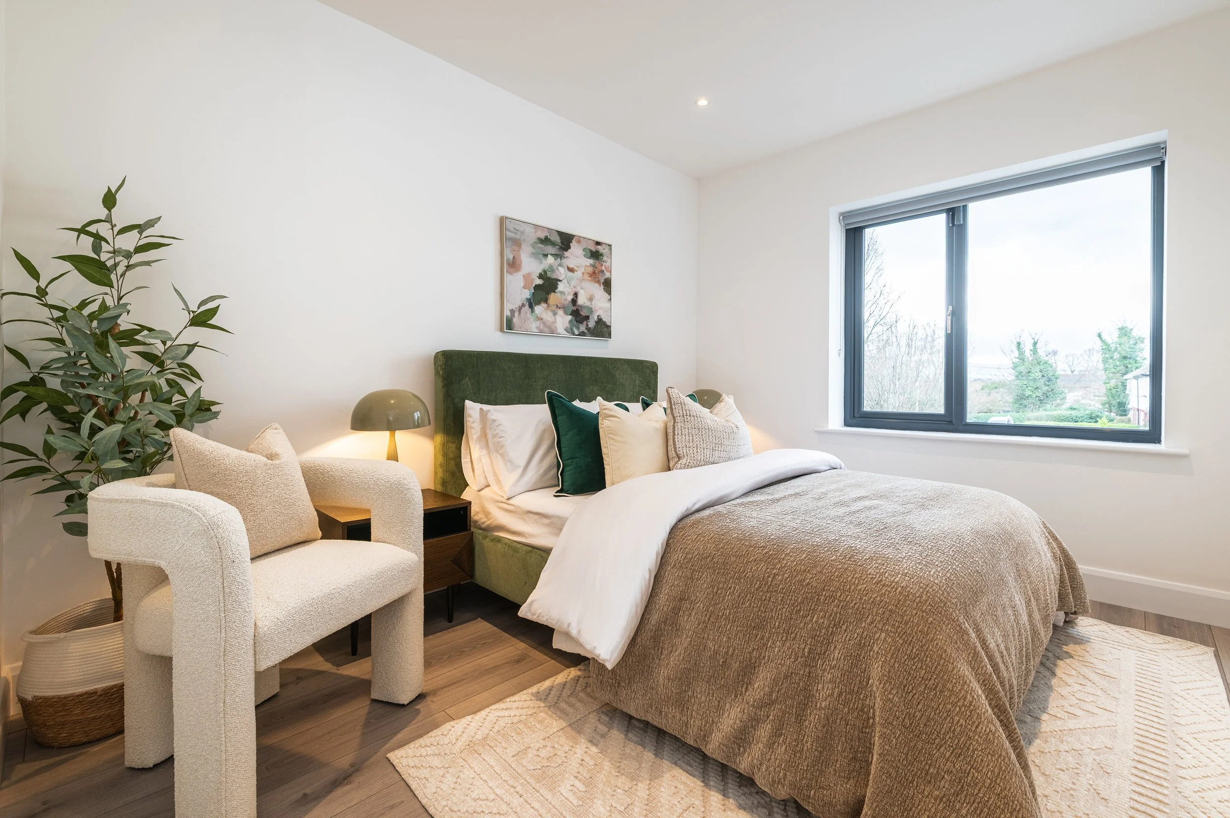 A cozy bedroom with a bed featuring a green velvet headboard, beige bedding, and decorative pillows. To the left, there's a white textured armchair with a beige cushion. A small bedside table holds a modern lamp with a greenish-gray shade. A large wi