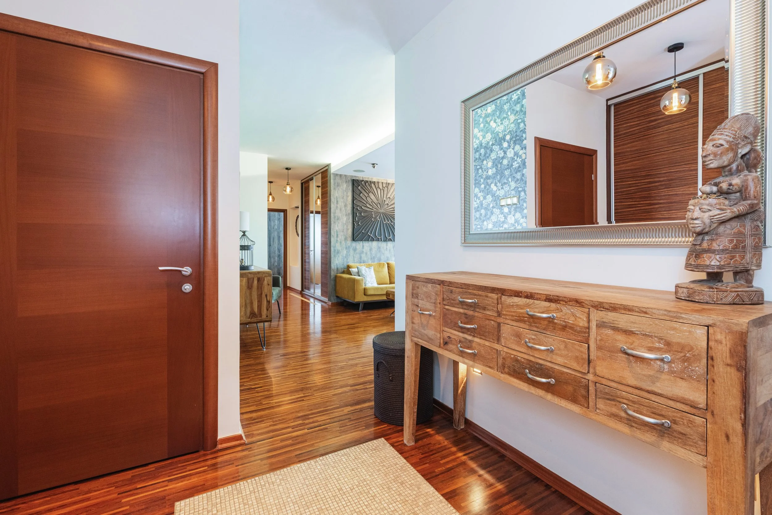 A modern interior hallway with wooden floors, a wooden dresser with drawers, a decorative wooden sculpture, a large mirror, and artwork on the walls. The hallway leads to a living area with a yellow sofa and various hanging light fixtures.