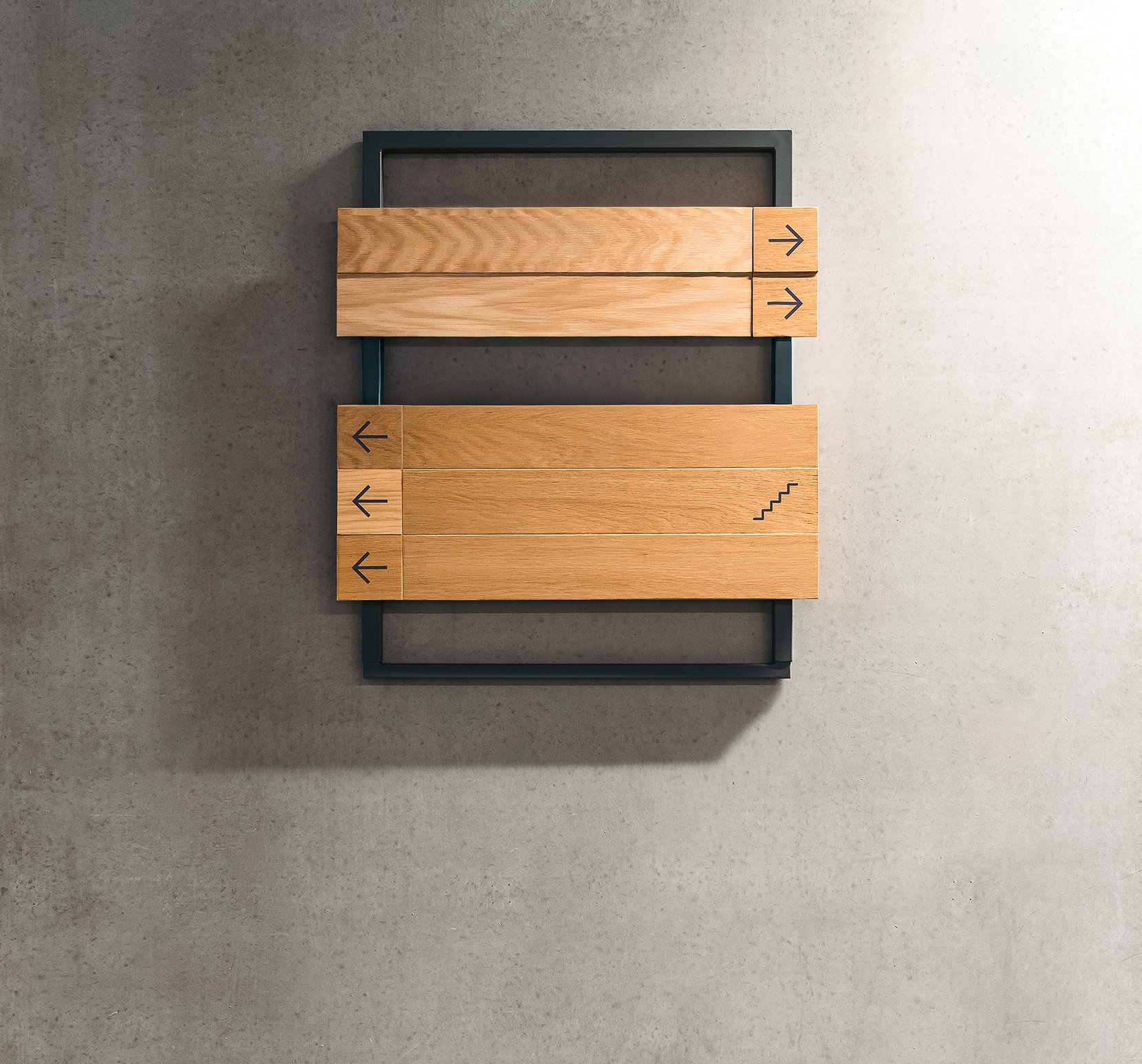 A modern piece of furniture with a black metal frame holding three wooden drawers with black arrows on the right and left sides, placed against a plain light-colored wall.