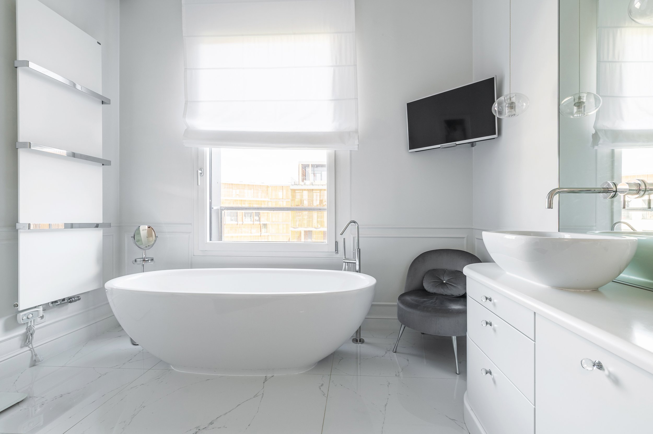 Modern white bathroom with a bathtub, a wall-mounted flat-screen TV, a sink with a large round bowl, a small black stool, and a window with white blinds.