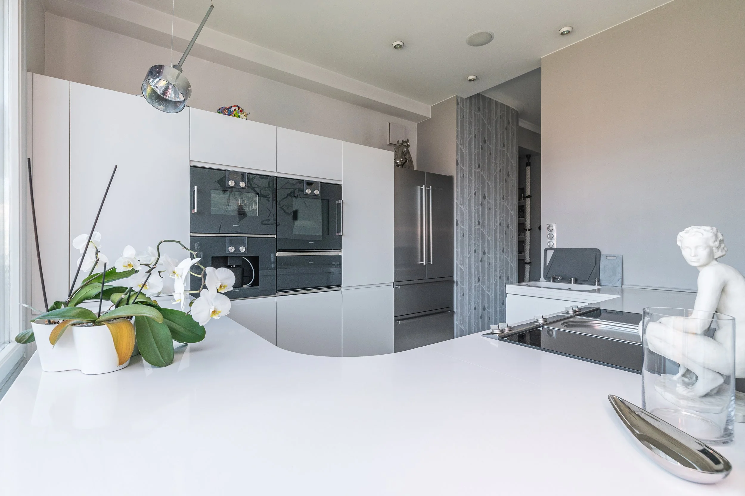 Modern kitchen with white and gray cabinets, a built-in oven and microwave, a stainless steel refrigerator, decorative items on top, a white counter with a potted orchid, and a sculpture of a woman. There is a gray barstool and a glass of water with 