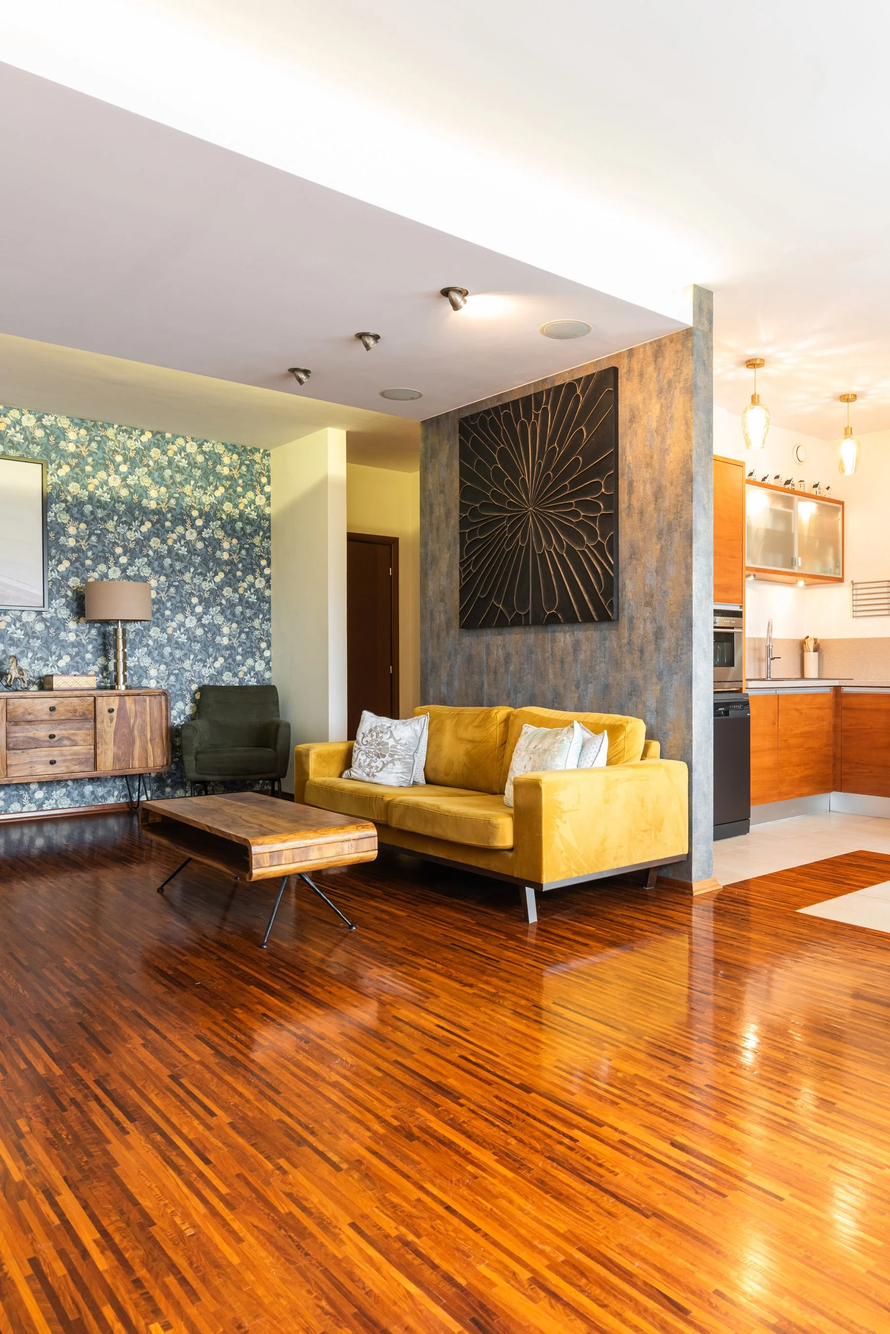 Living room with yellow sofa, wooden coffee table, floral wallpaper, and artwork on the wall. Open kitchen with hanging lights, cabinets, and appliances. Wooden flooring throughout.
