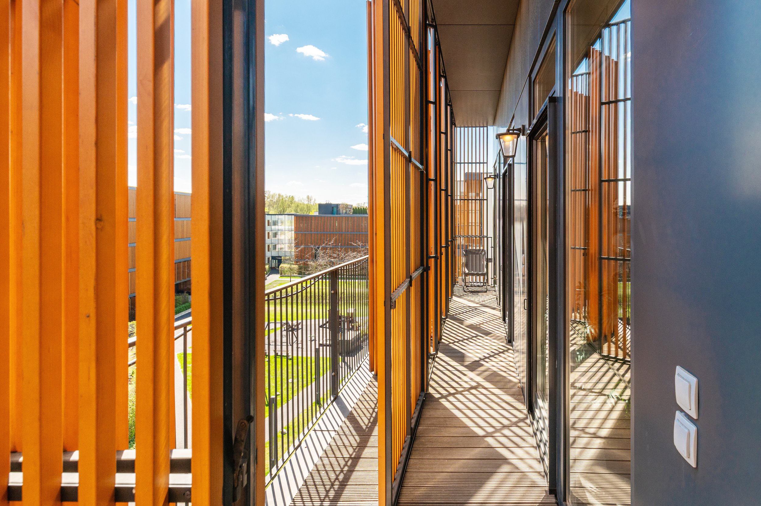 View from a balcony showing wooden slats on the left, glass doors on the right, warm outdoor lighting, and a distant view of buildings and trees under a blue sky with some clouds.