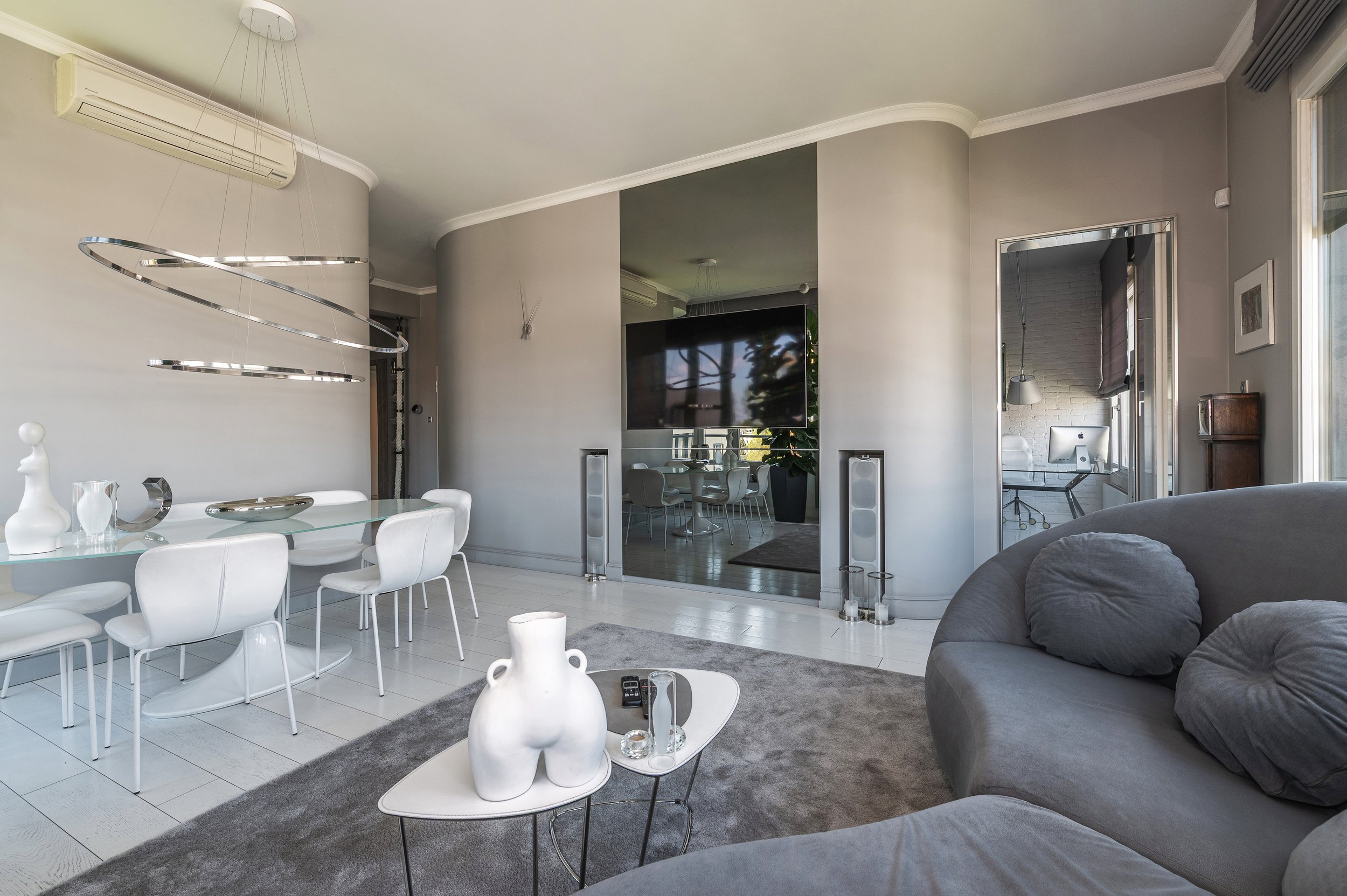 A modern living room with a grey sofa, a round white coffee table, and a dining area with white chairs and a glass dining table. There is a large flat-screen TV, a potted plant, and a doorway leading to a home office with an iMac computer and a hangi
