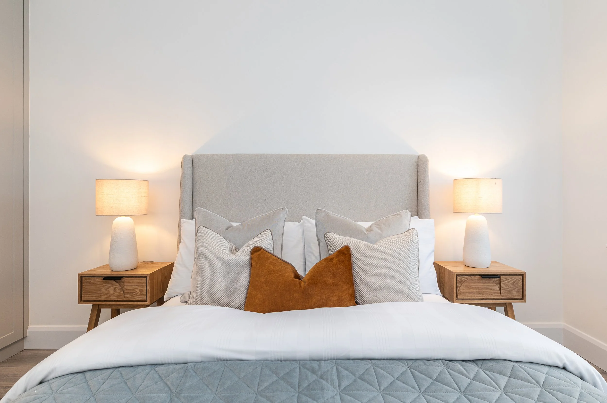 A neatly made bed with a light-colored upholstered headboard, multiple pillows including a large rust-colored pillow, and two wooden nightstands with lamps on either side, in a minimalist bedroom with a plain white wall.