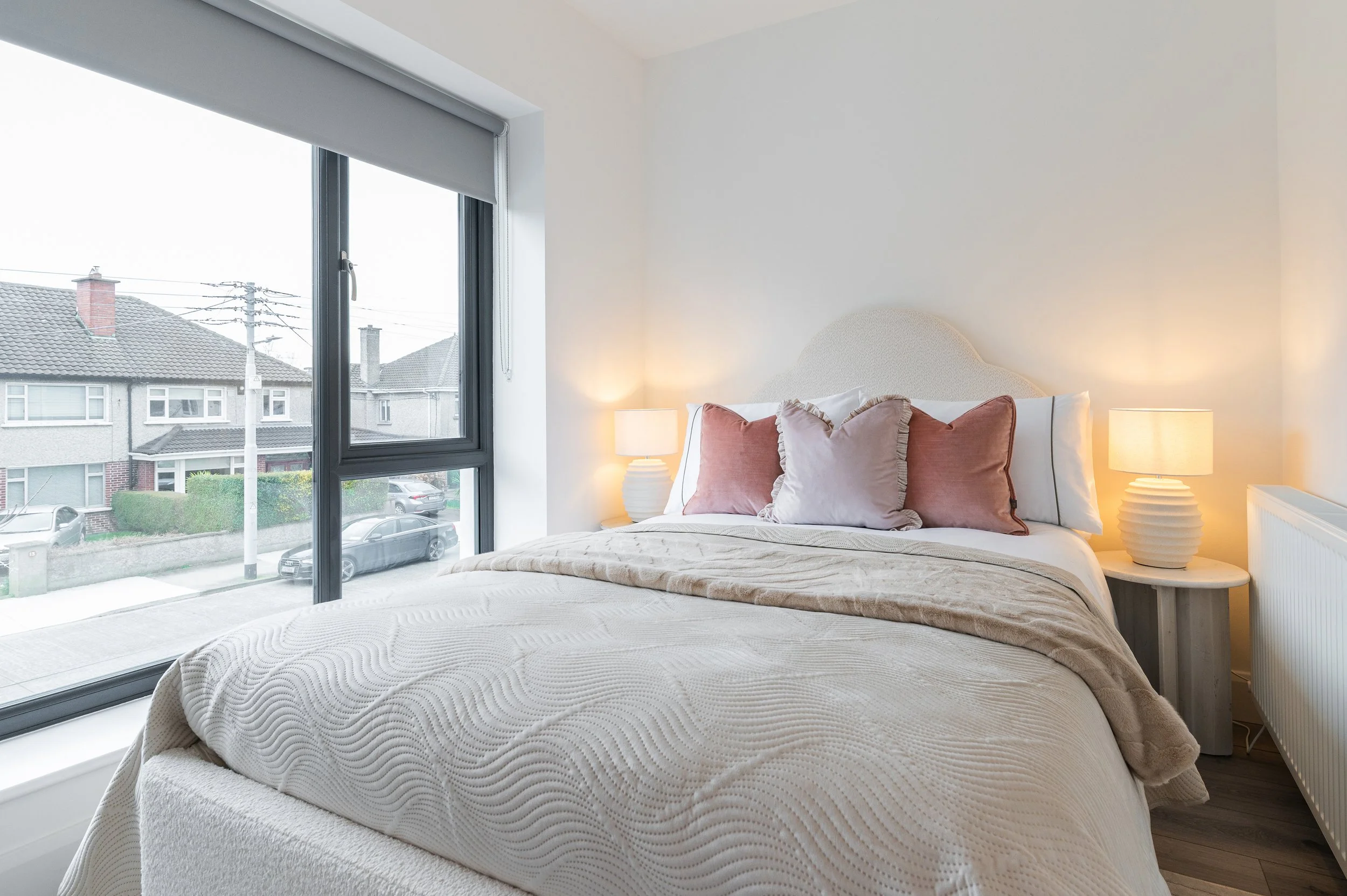 A bedroom with a large window showing a city street view, a bed with beige and pink pillows, and two lamps on nightstands.